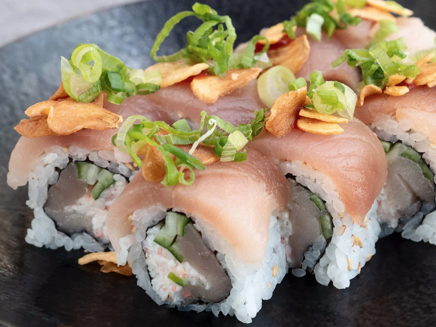 Close-up of sushi rolls topped with sliced green onions and crispy garlic chips, on a black plate.