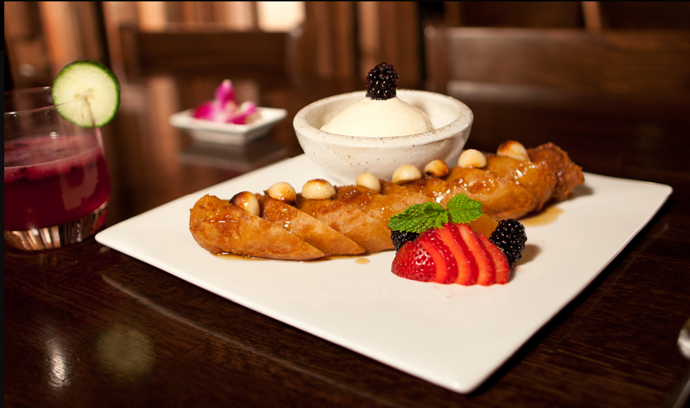 A plate with fried banana drizzled with syrup, topped with vanilla ice cream and blackberries, garnished with strawberry slices and mint leaves, served with a glass of berry juice garnished with cucumber, and a small dish of pink pickled vegetables i