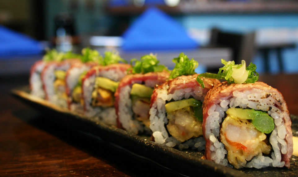 Close-up of a platter of sushi rolls garnished with green onions, with a blurred restaurant background.