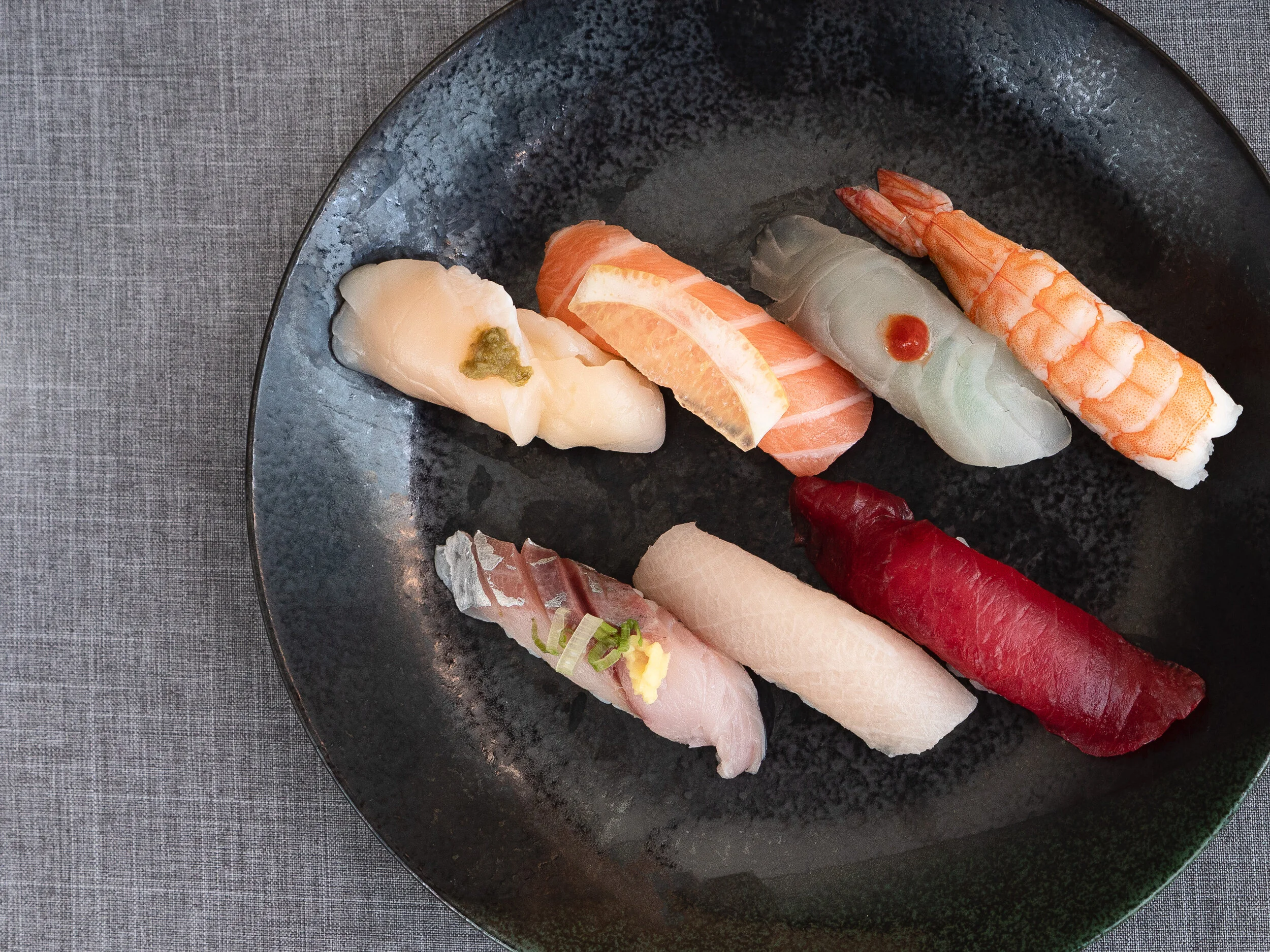 A black round plate with an assortment of eight pieces of sushi on a gray textured surface.