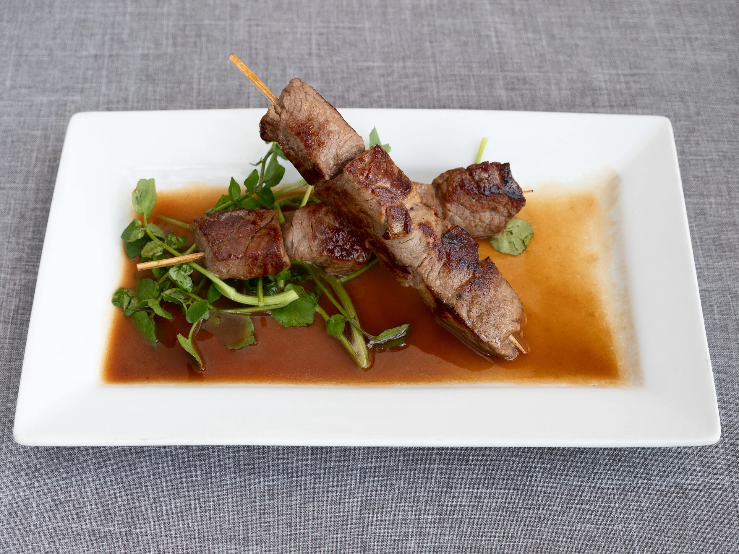Grilled beef skewers on a bed of fresh greens with set in a brown sauce on a white rectangular plate.