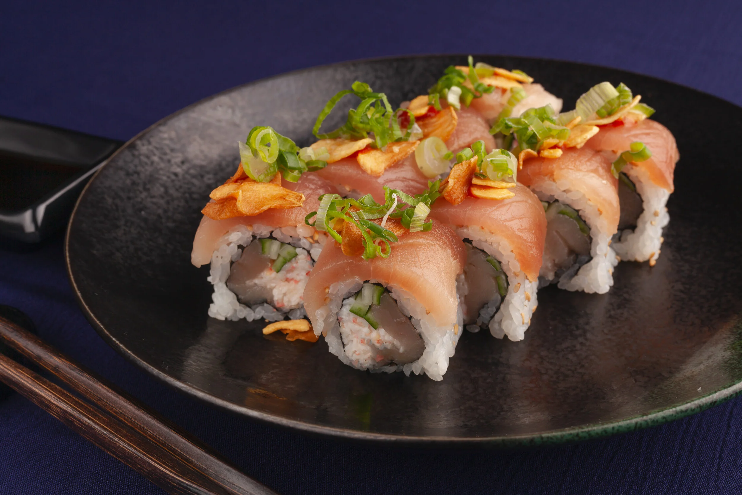 Sushi rolls topped with sliced fish, green onions, and crispy garlic on a black plate.