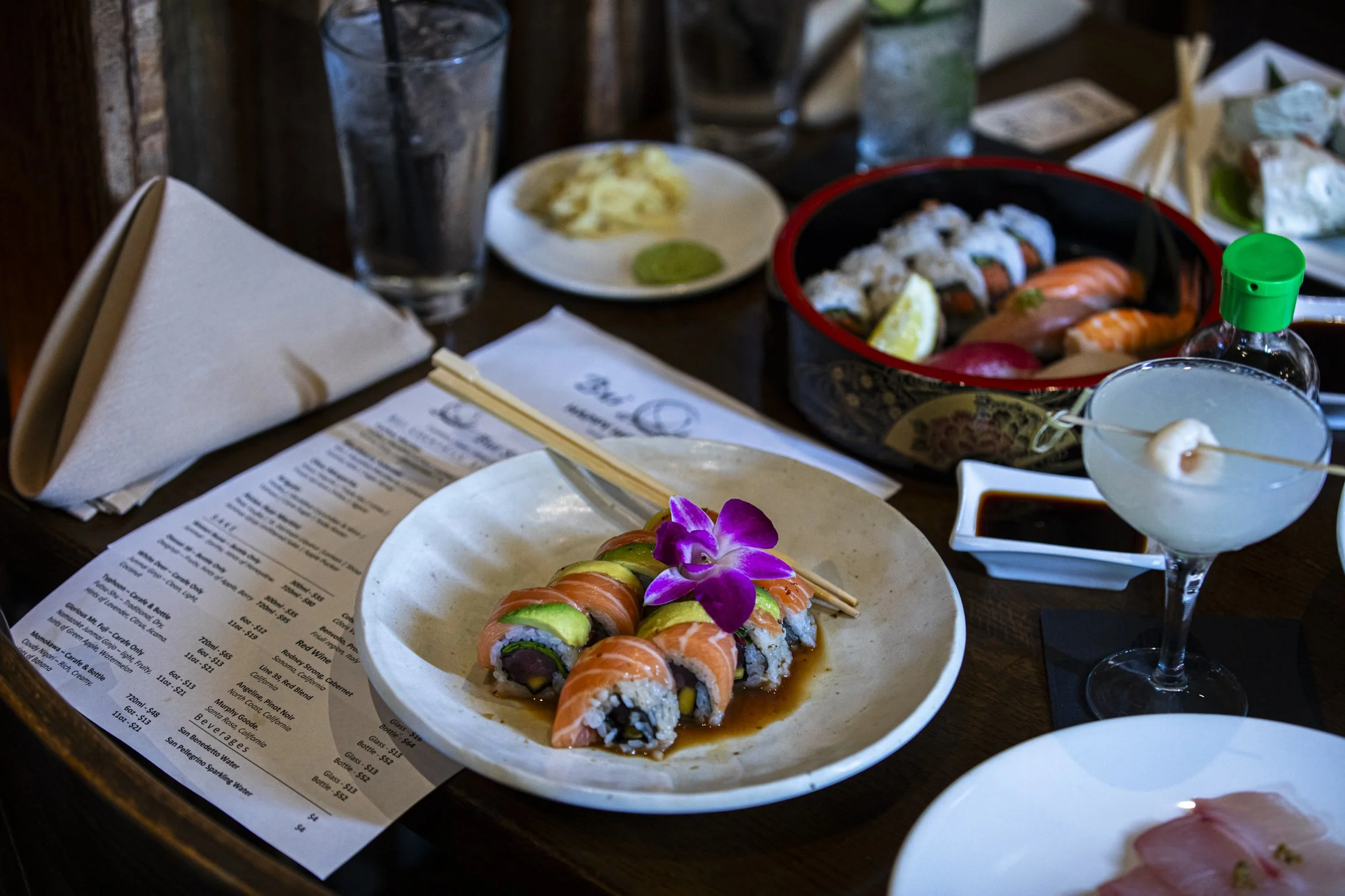 Close-up of a table setting with sushi rolls garnished with an edible flower, a menu, and various Asian dishes, drinks, chopsticks, and condiments visible.
