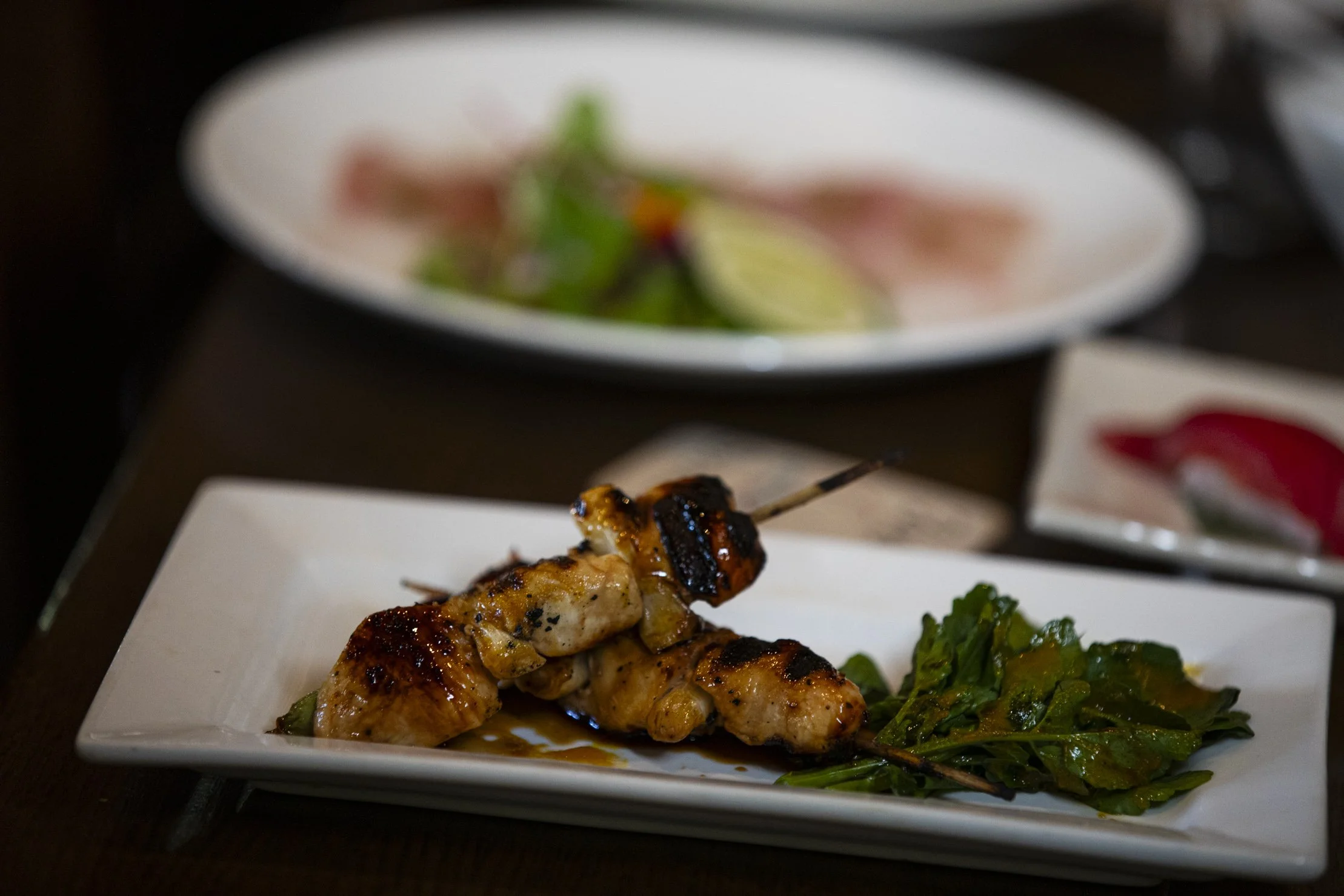 Grilled chicken skewer on white rectangular plate with greens, blurred plates in background
