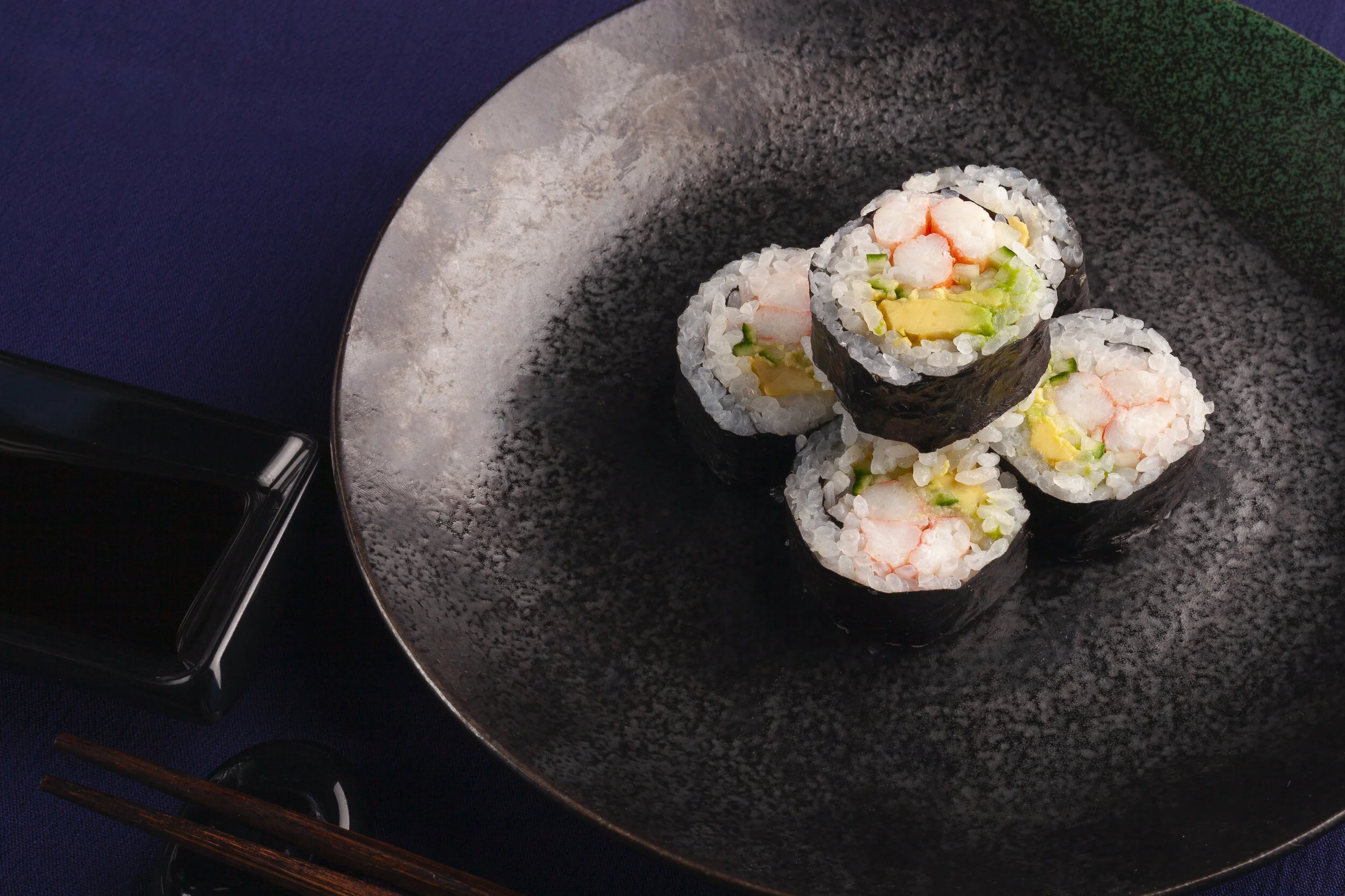 Five pieces of sushi with shrimp, avocado, and cucumber in rice and seaweed, arranged on a black textured plate.