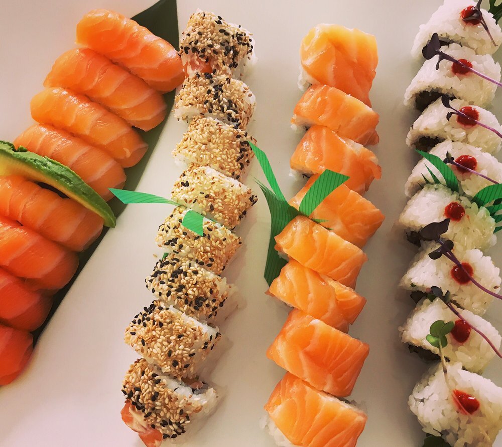Assorted sushi rolls arranged on a white plate, including salmon sashimi with lime wedge, sesame seed-covered rolls, salmon nigiri, and white rice rolls topped with sauce and herbs.