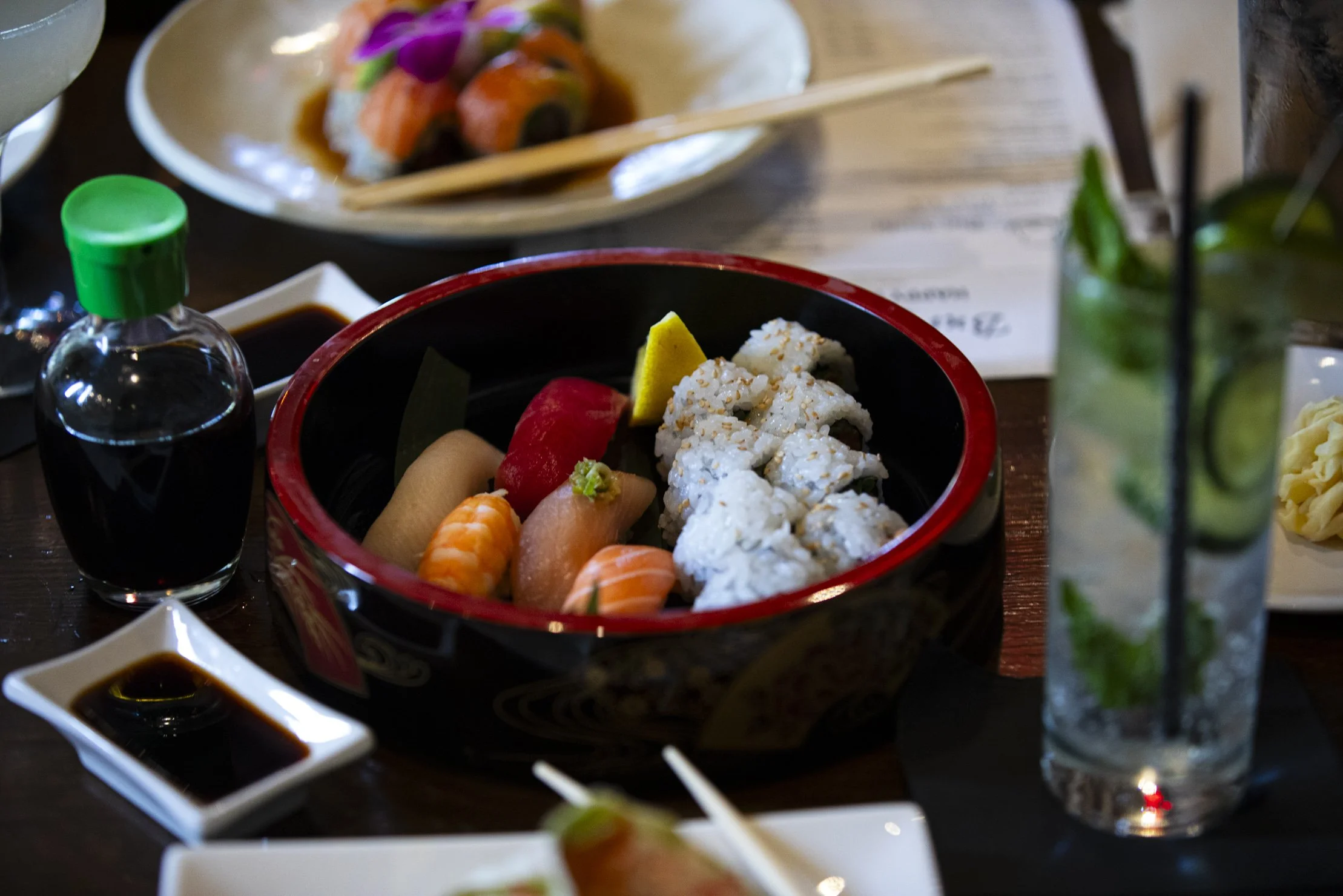 A bowl of assorted sushi including sashimi, rolls with sesame seeds, and garnishes like lemon and wasabi on a table with soy sauce, a bottle of soy, a glass of mojito with mint and cucumber, and other dishes.