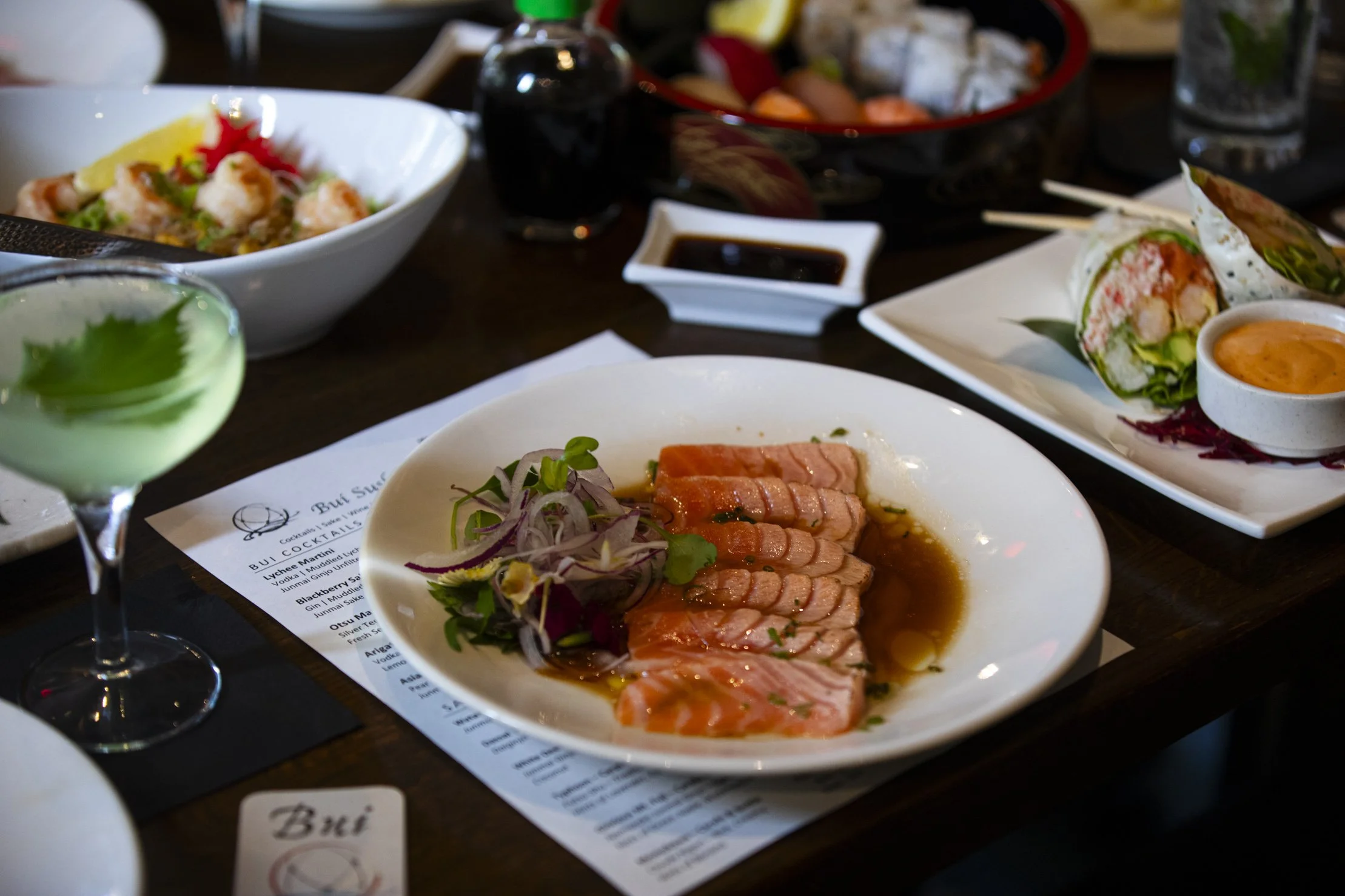 Assorted Japanese dishes on a dark wooden table, including sashimi with onions, mini rolls with sauce, a green cocktail, and other bowls of food.