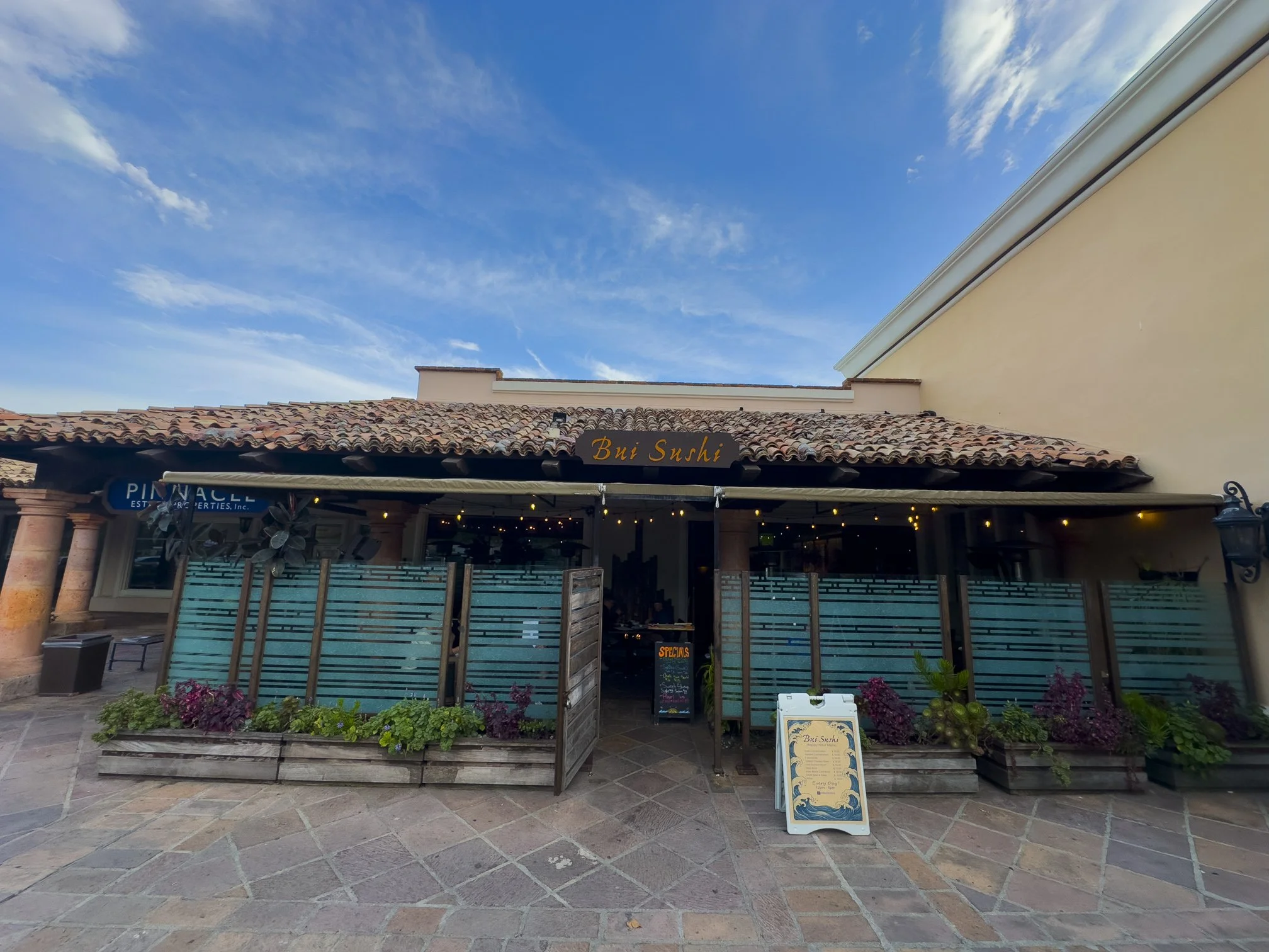 Exterior of a restaurant named Bui Sushi with a tiled roof, decorative plants in front, and a signboard outside on a paved area under a blue sky.