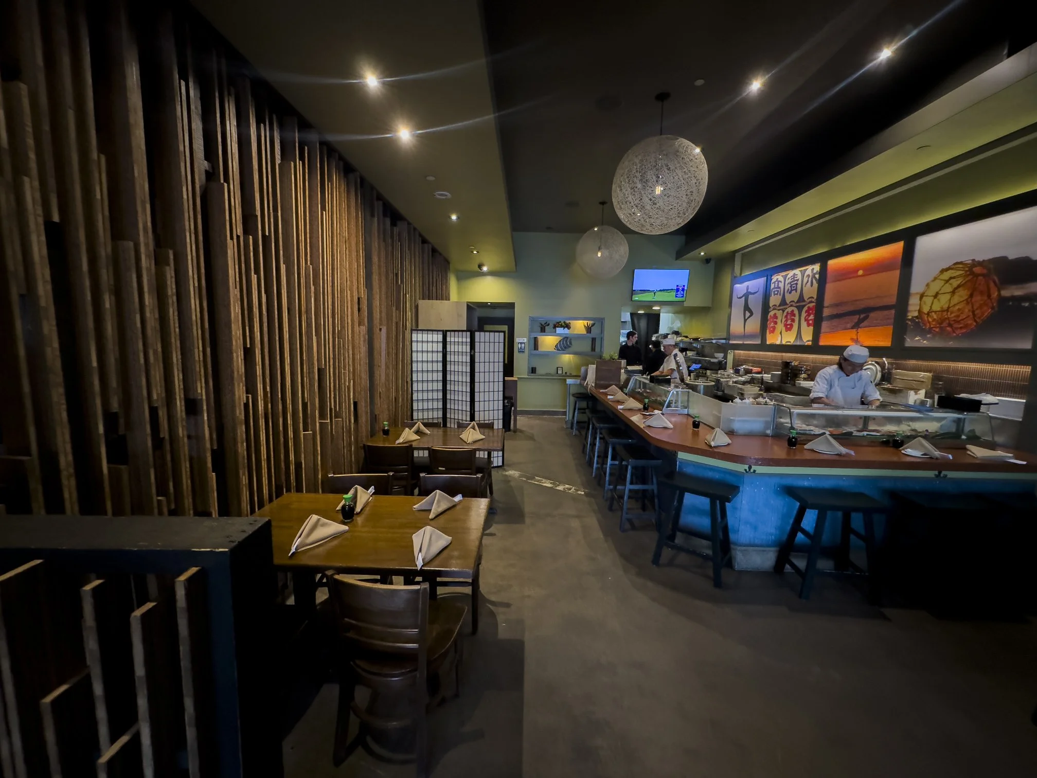 Interior of a Japanese restaurant with a bar area, Korean chefs working, tables set with folded napkins and soy sauce bottles, wooden wall decor, television screens, and Japanese-themed artwork.