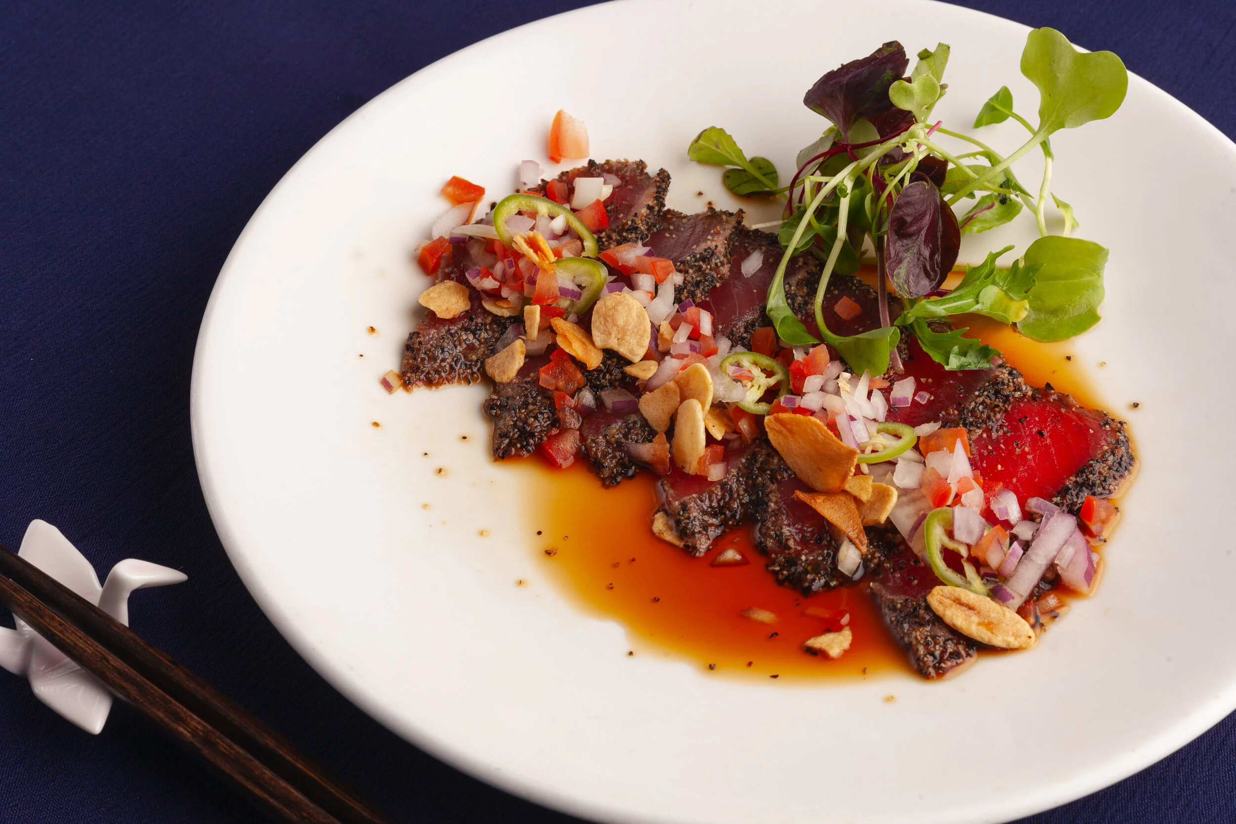 Seared tuna sashimi with chopped red onions, sliced green chili peppers, crushed peanuts, fresh microgreens, and a soy-based sauce on a white oval plate.