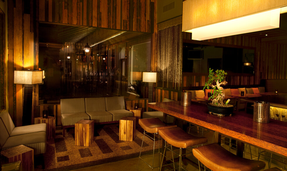 Elegant modern lounge with wooden furniture, couches, bar seating, and a potted plant on the table, illuminated by warm lighting.