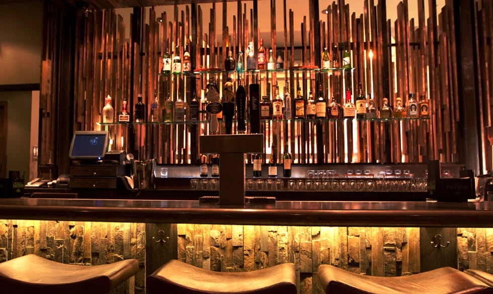 A bar with a backlit display of various liquor bottles on glass shelves, a countertop with a cash register, and bar stools in the foreground. The background has a decorative wooden wall.