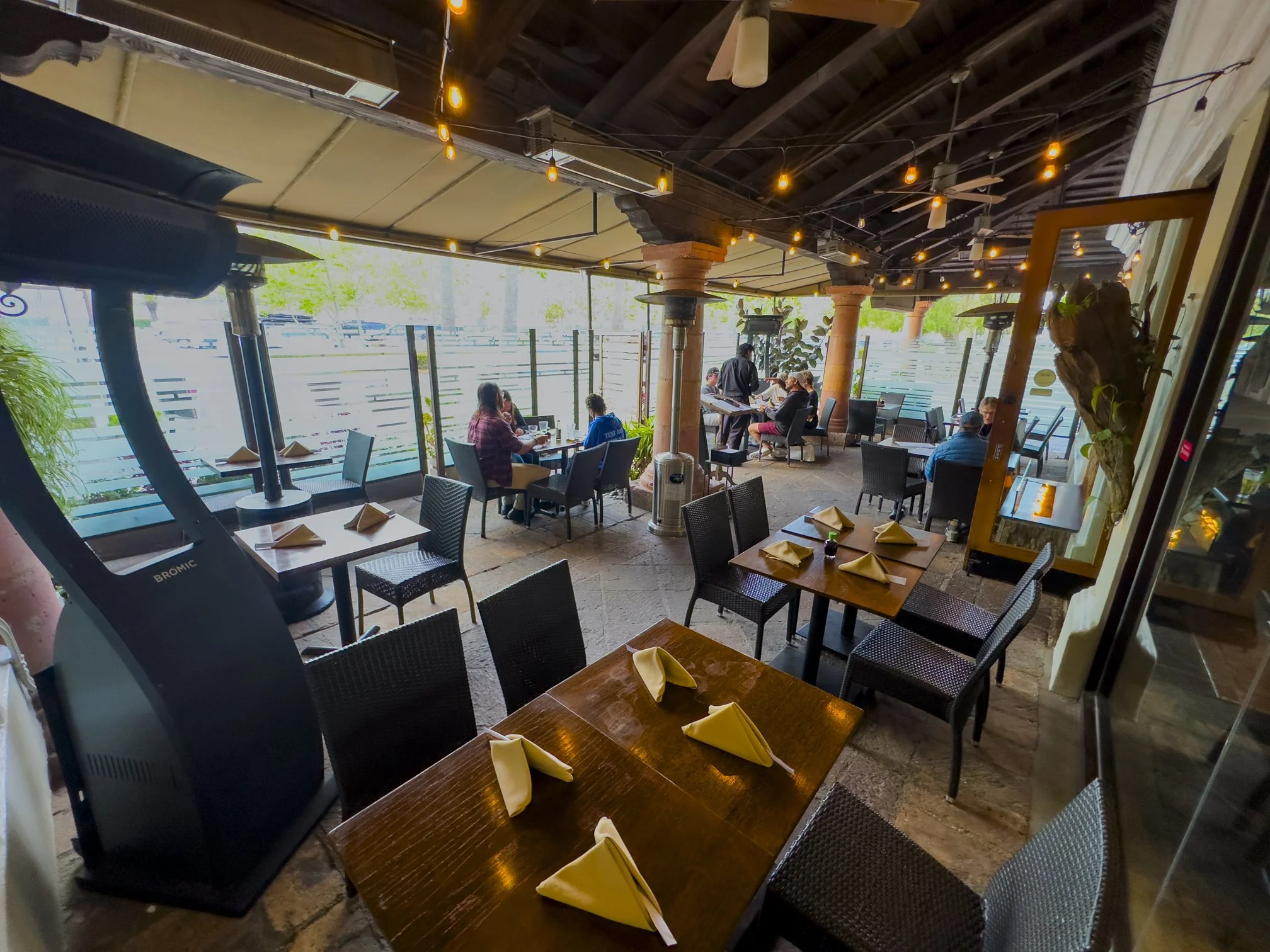 Interior view of a restaurant with outdoor seating, tables set with napkins, and a few customers dining.