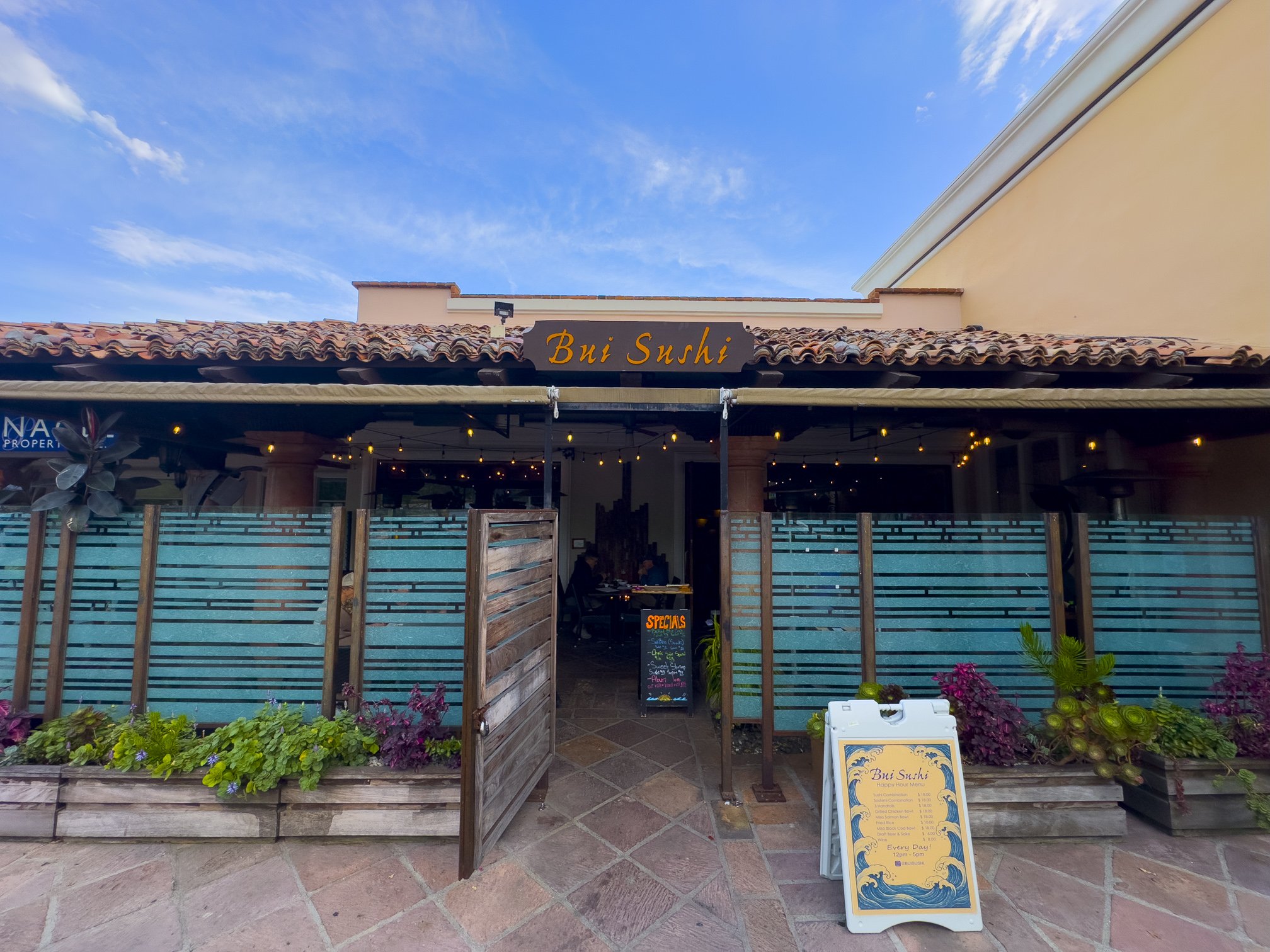 Exterior of Bui Sushi restaurant with an open wooden gate, a decorative sign, and a chalkboard menu outside, surrounded by potted plants and blue fencing under a clear sky.