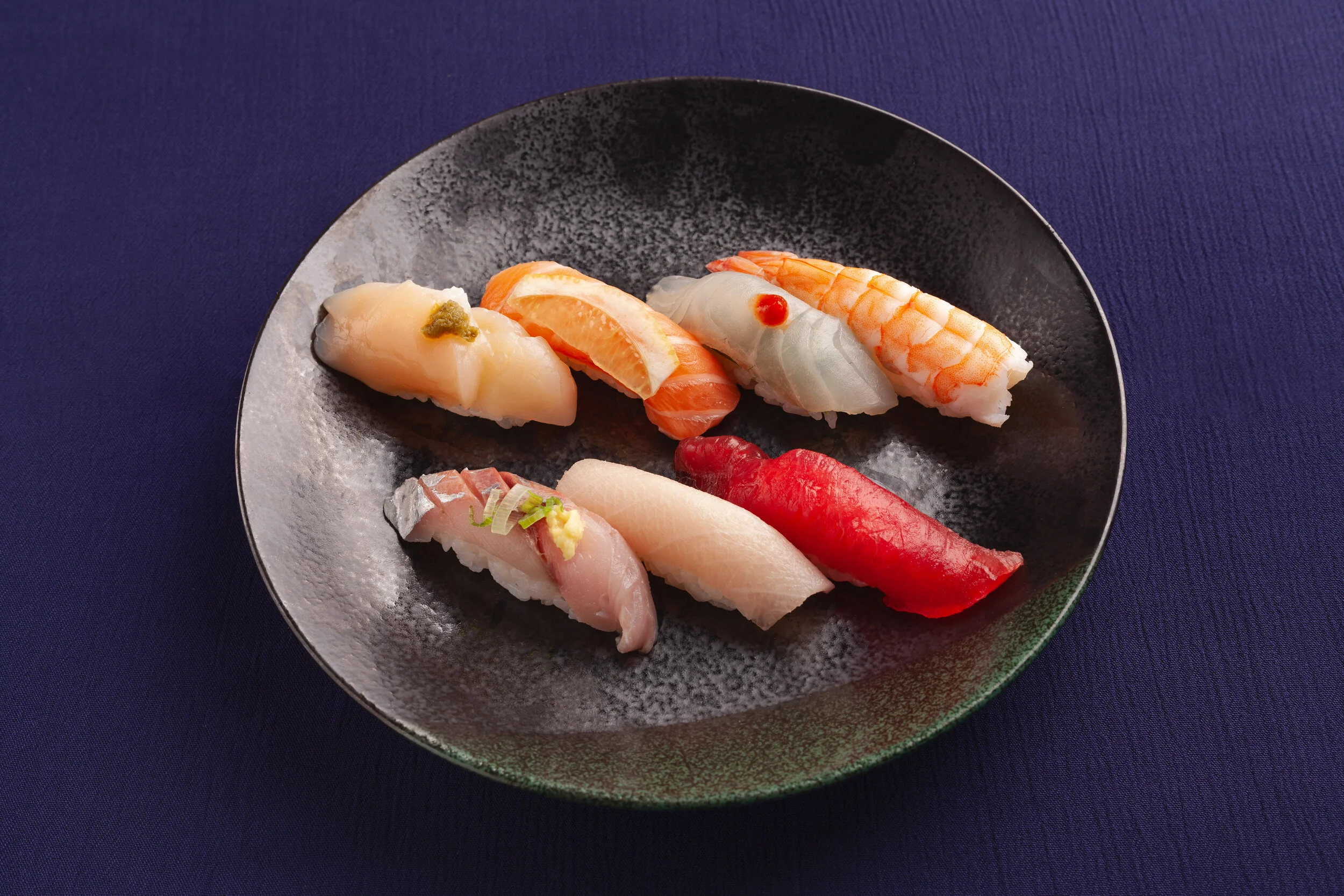 Assorted sushi pieces on a black plate against a dark blue background.