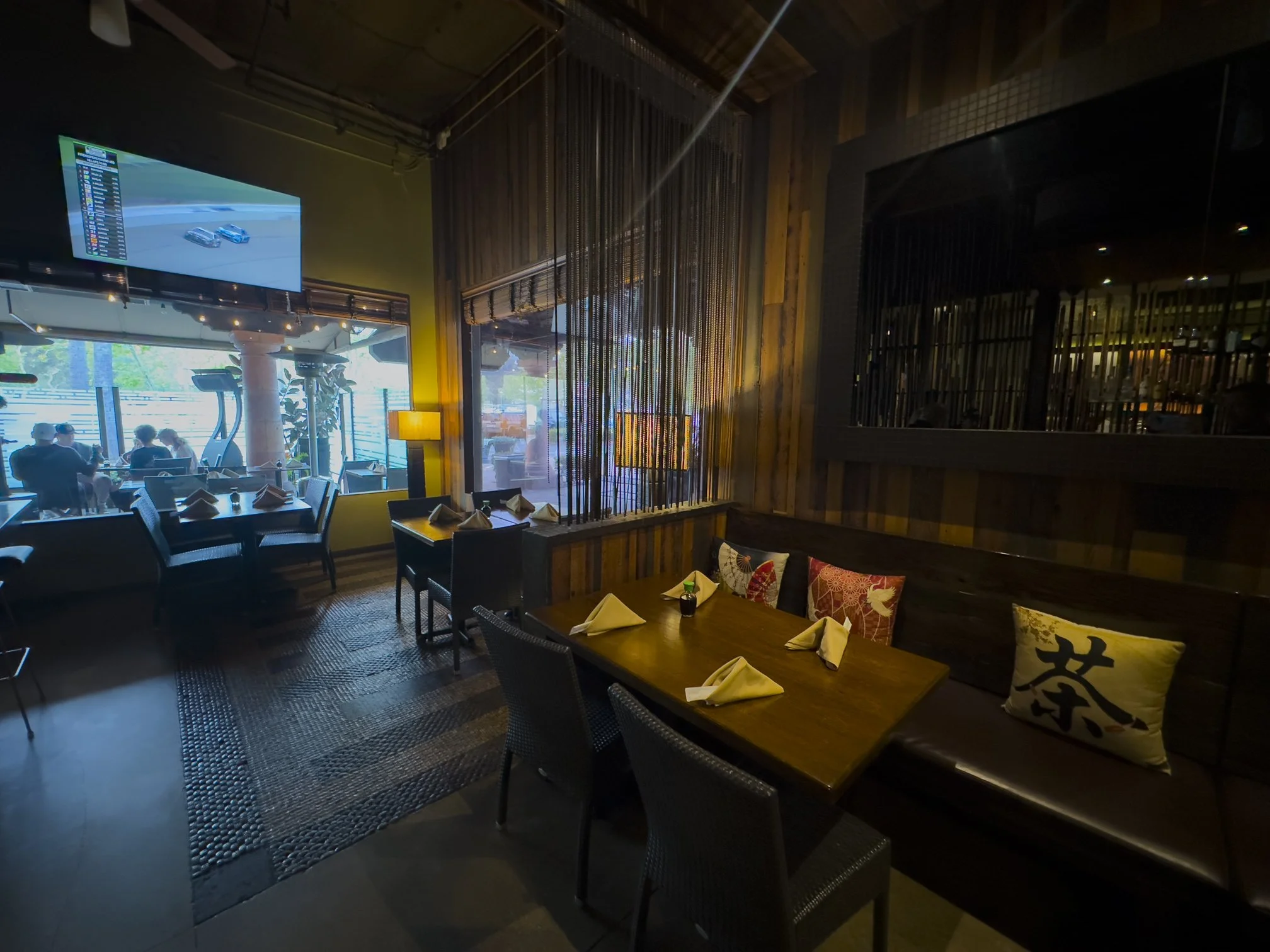 Inside a dimly lit restaurant with a wooden interior, a table set with napkins and condiments, decorative pillows on the bench, and a large window showing outdoor seating and a TV screen.