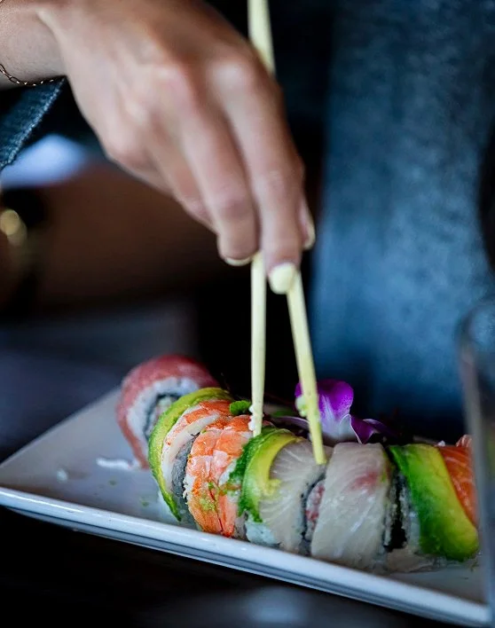 Person using chopsticks to pick up sushi rolls on a white rectangular plate, with colorful fish and cucumber on the sushi.