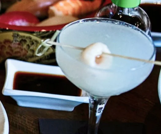 A cocktail glass with a frozen drink, garnished with a shrimp on a skewer, placed on a dark table with a dipping sauce and other condiments in the background.