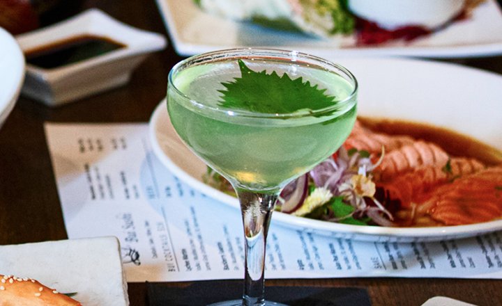 A cocktail glass with green liquid and a leaf garnish on top, placed on a table with Asian food dishes.