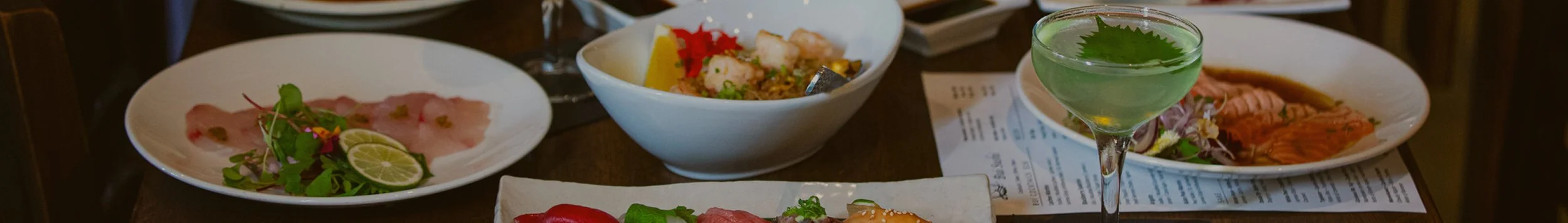 Various dishes and a green cocktail on a restaurant table, including thinly sliced carpaccio, a seafood bowl, and slices of smoked salmon.