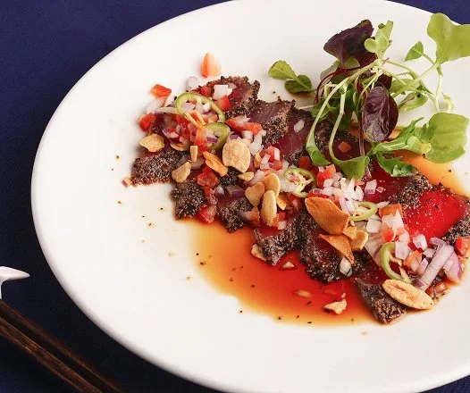 A white plate with a dish featuring sliced raw meat, chopped onions, tomatoes, and green onions, garnished with microgreens, chopped peanuts, and a drizzle of sauce.