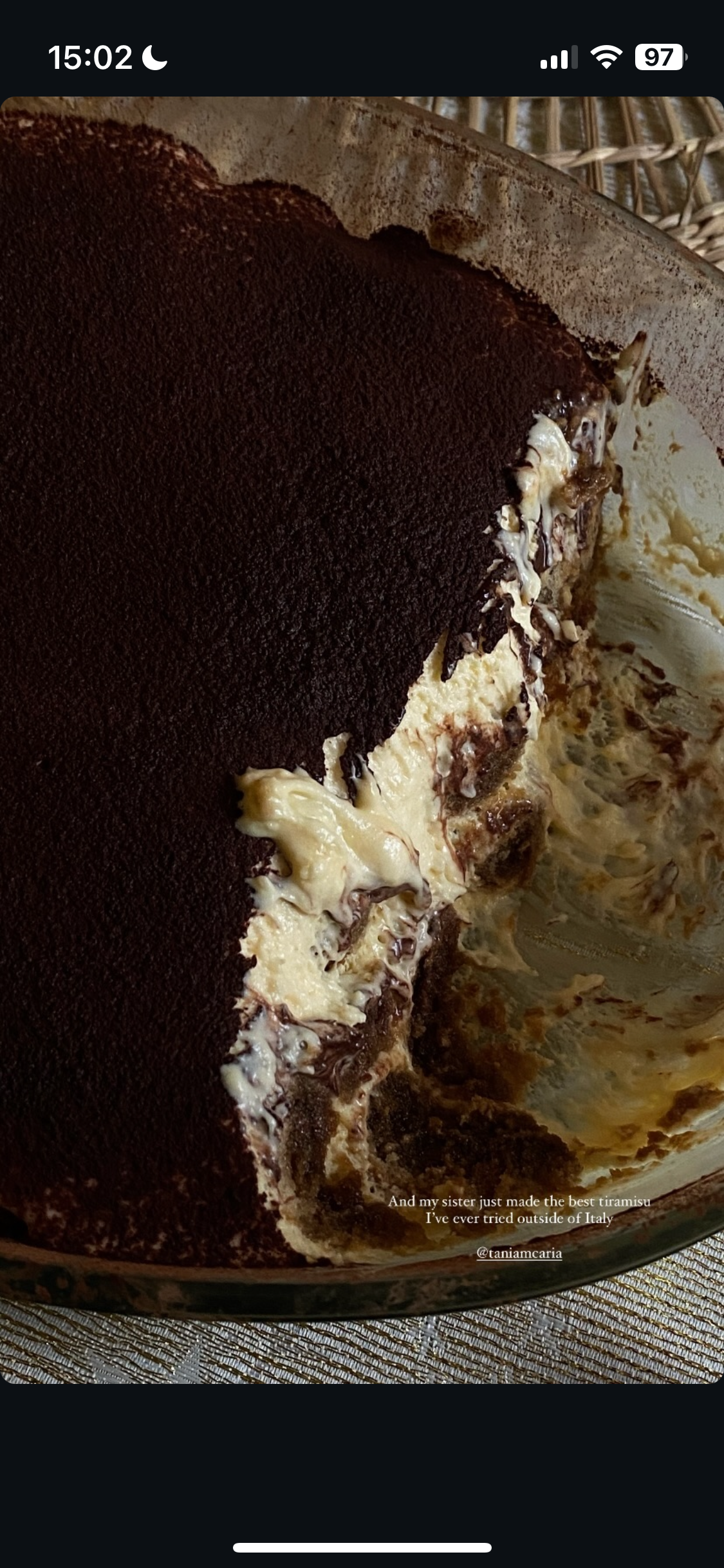 A half-eaten tiramisu in a glass dish with cocoa powder on top, showing layers of mascarpone cheese and coffee-soaked ladyfingers.