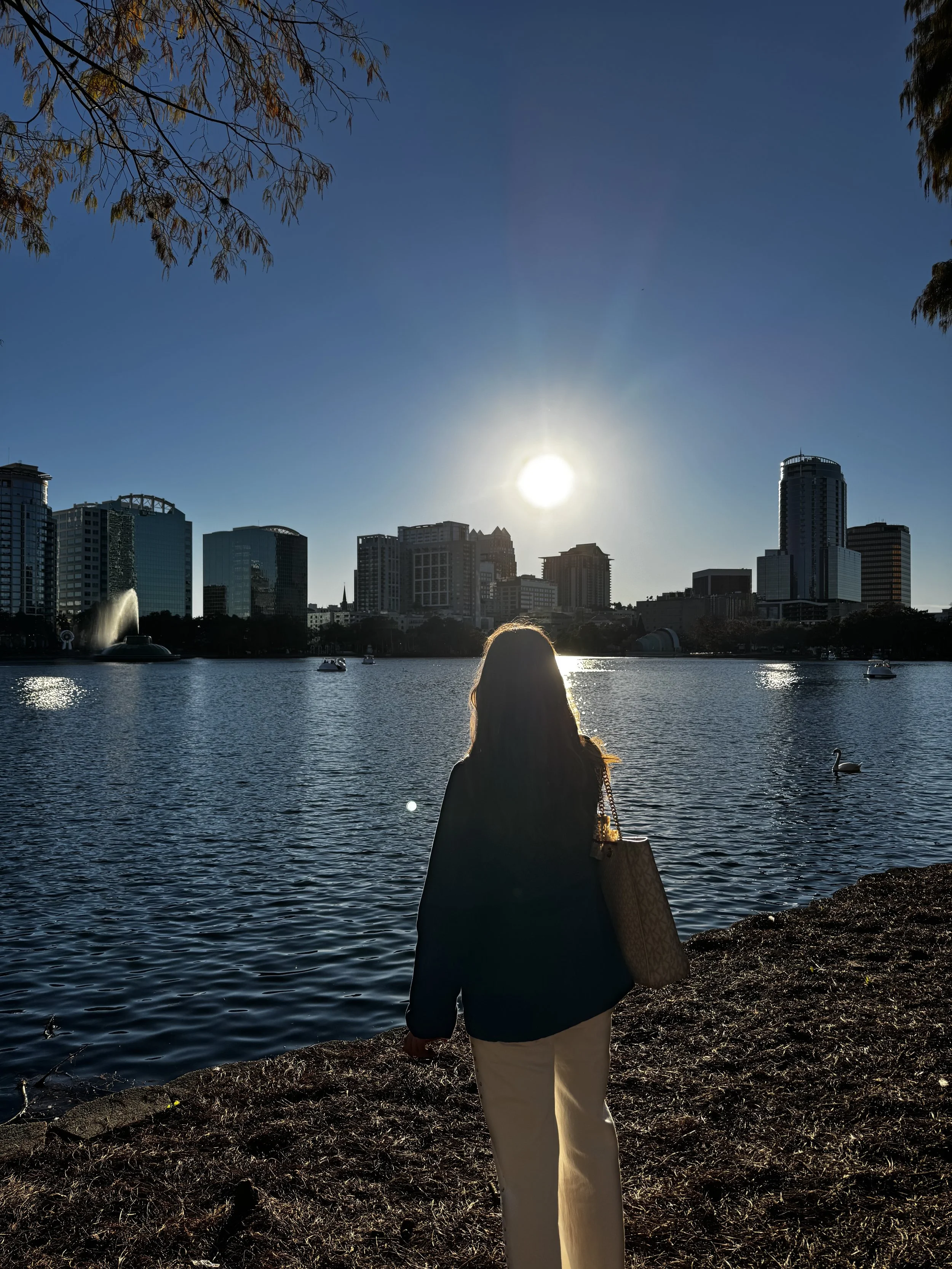 A person standing by a body of water with a city skyline in the background during sunset.