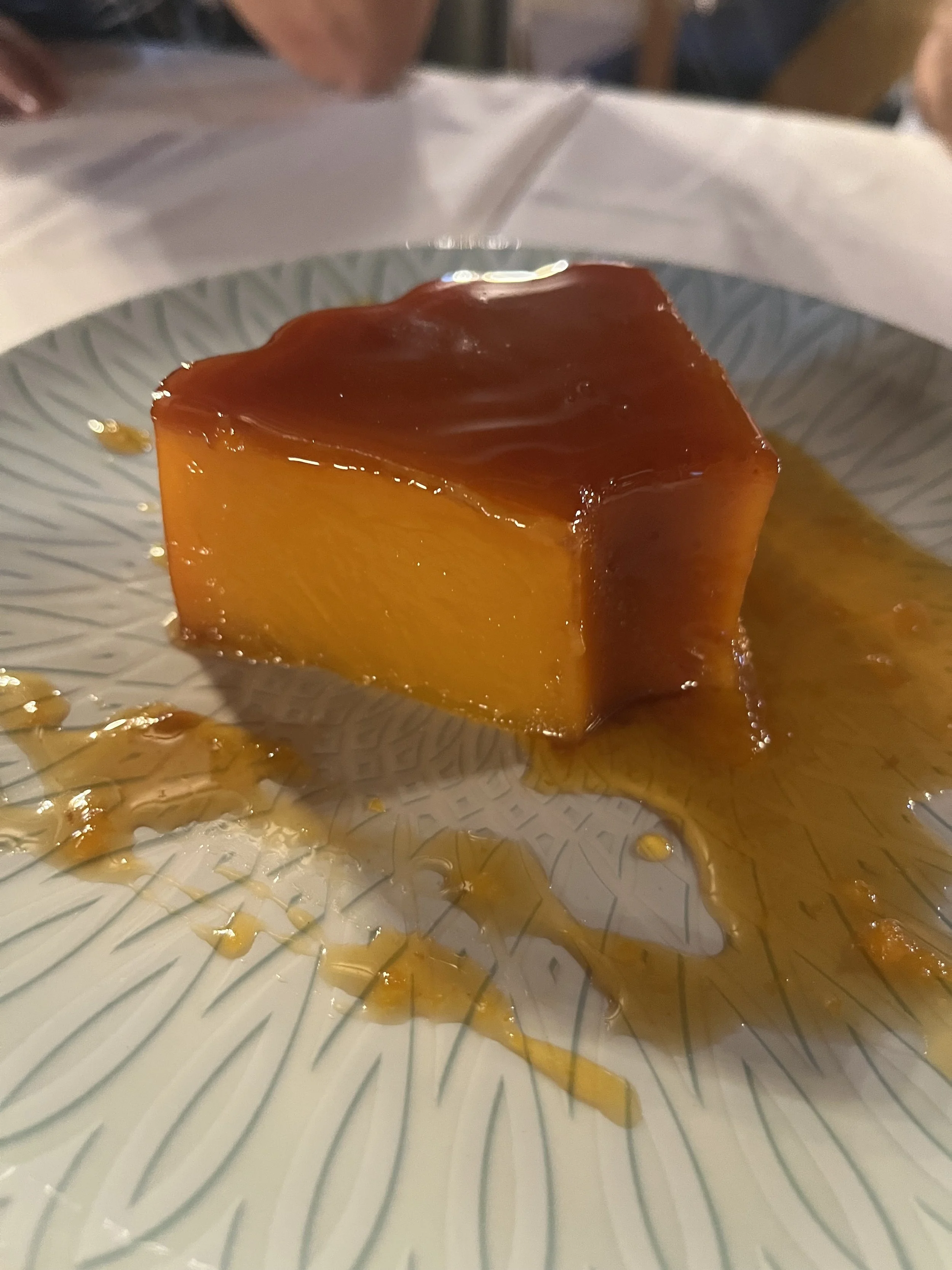 A slice of flan with caramel sauce on top, placed on a patterned plate with some caramel sauce around it.