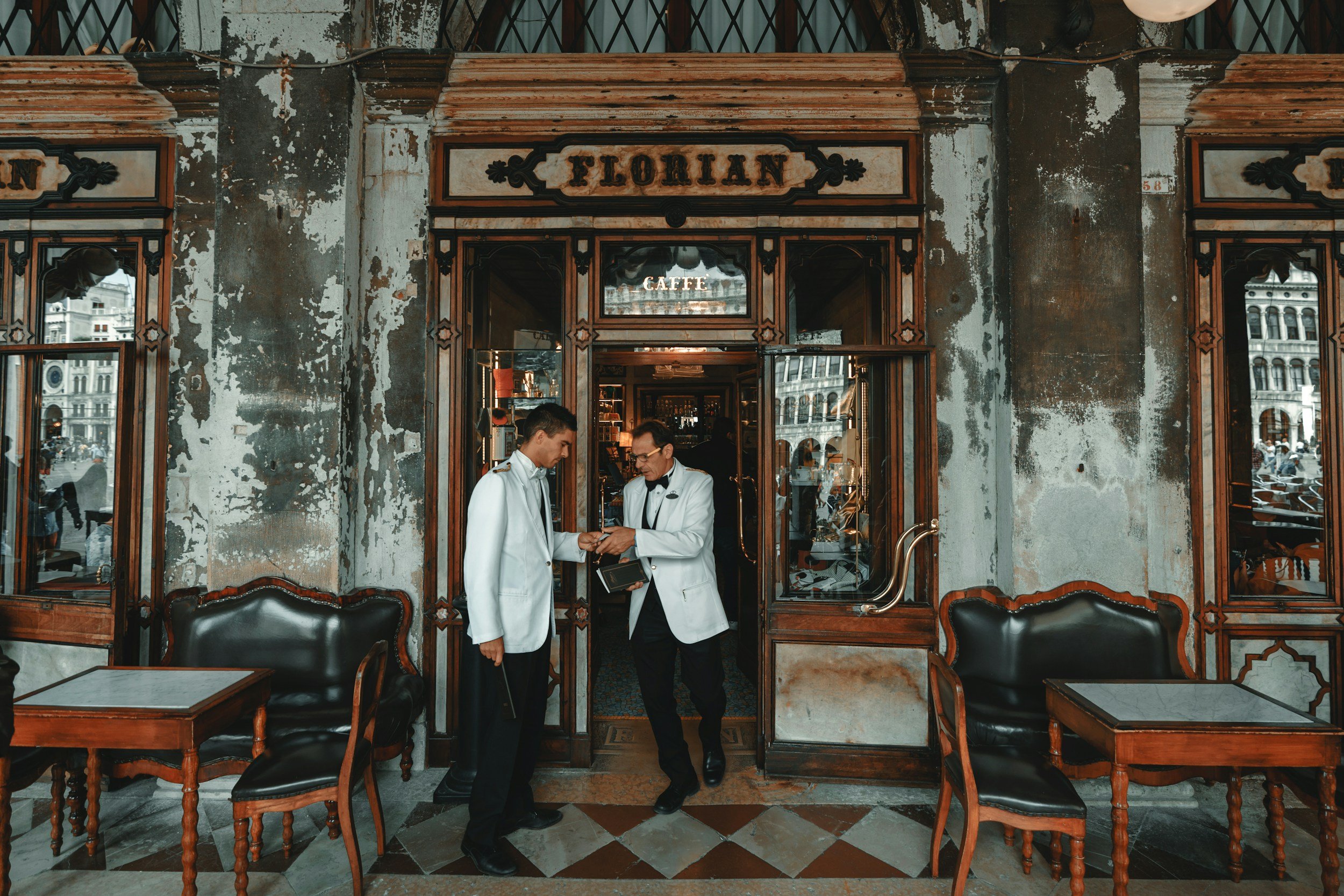 Two caterers dressed in white uniforms standing in front of an old restaurant entrance, with reflective glass doors labeled 'Floridan'. The restaurant has a vintage, worn exterior with peeling paint and ornate wooden framing. There are black leather chairs and small tables outside, and the scene appears to be in a busy city.