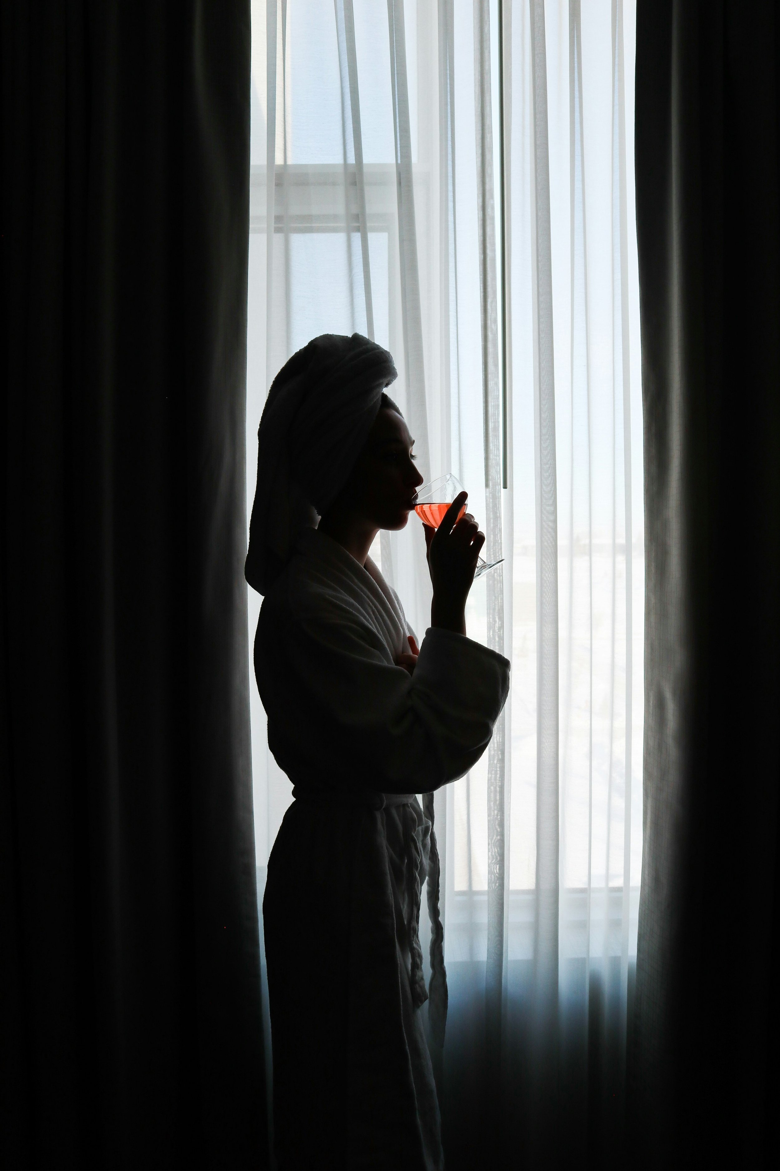Silhouette of a woman in a bathrobe and towel on her head, drinking from a glass with pink drink, standing beside a window with sheer curtains.