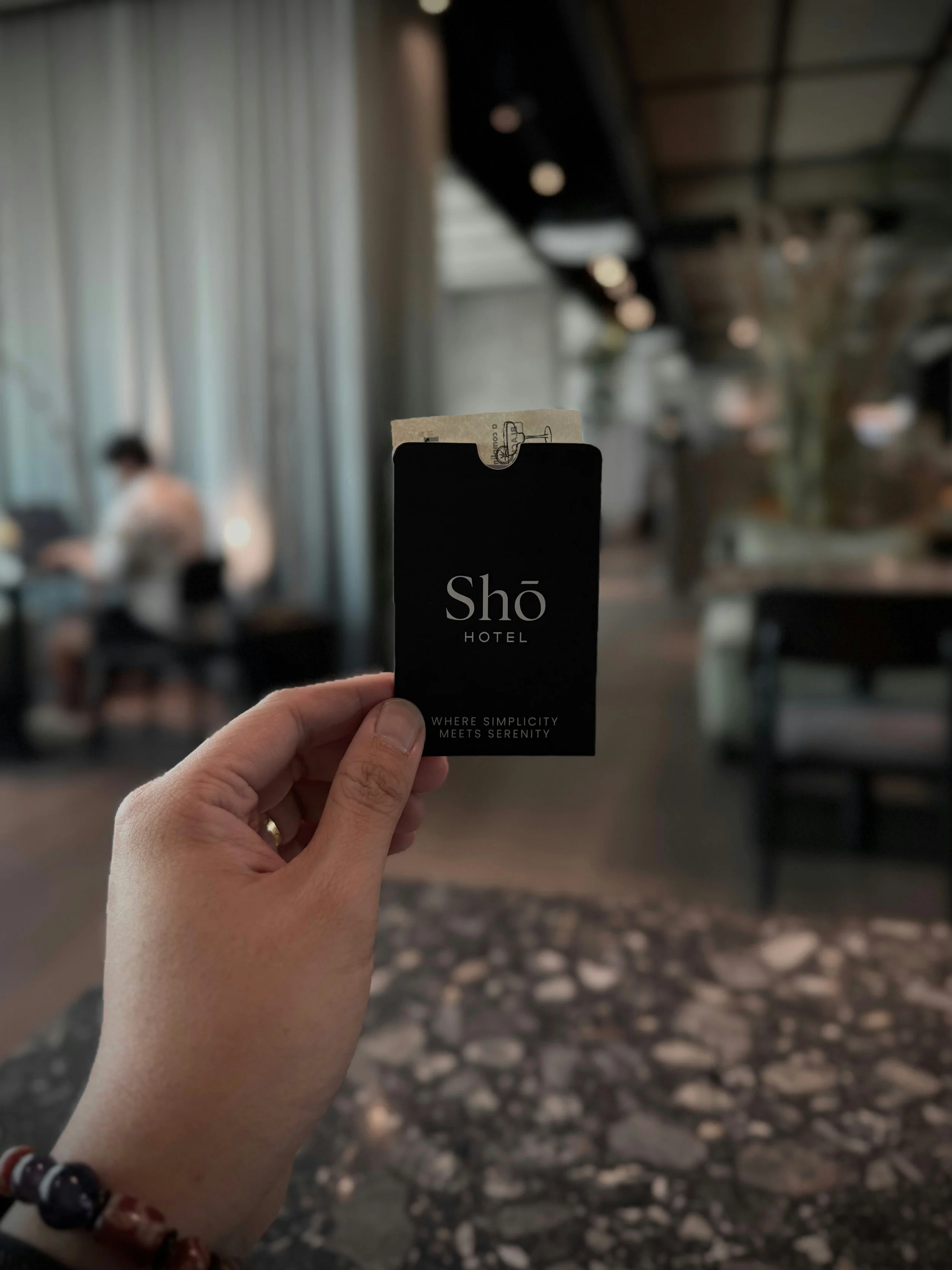 Person holding a black key card for Shō Hotel, with a tag sticking out, inside a modern lobby with blurred background of chairs and large windows.