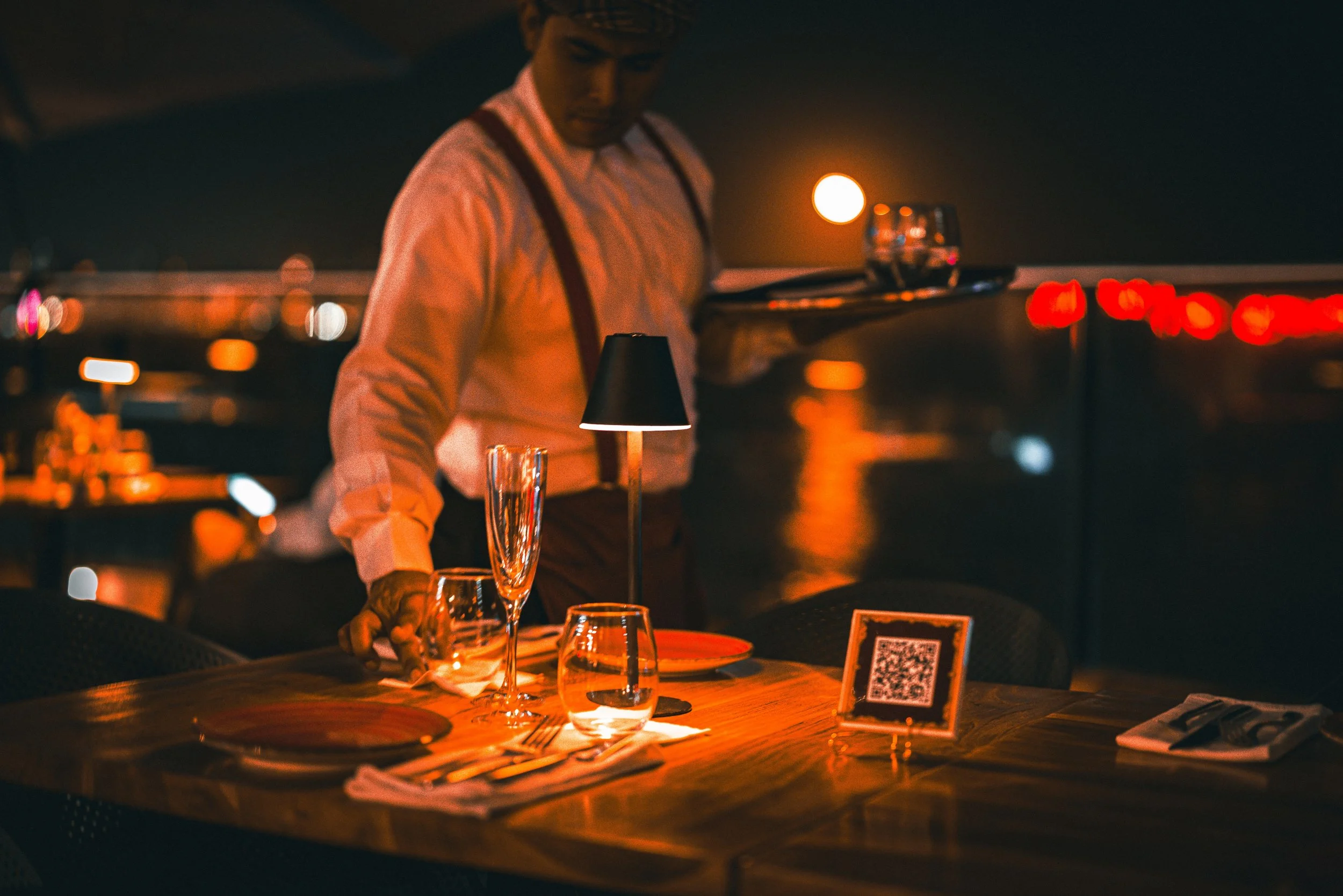 A waiter arranges glasses and plates on a dimly lit restaurant table near a waterfront at night.
