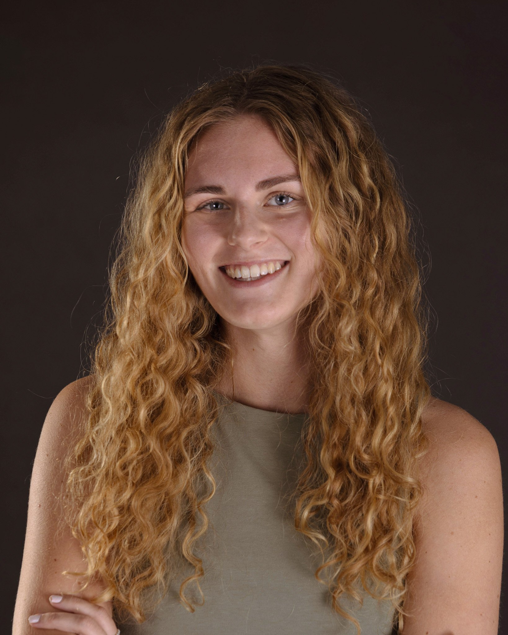 Eine junge Frau mit lang, lockigem, blondem Haar, die lächelnd vor dunklem Hintergrund posiert.