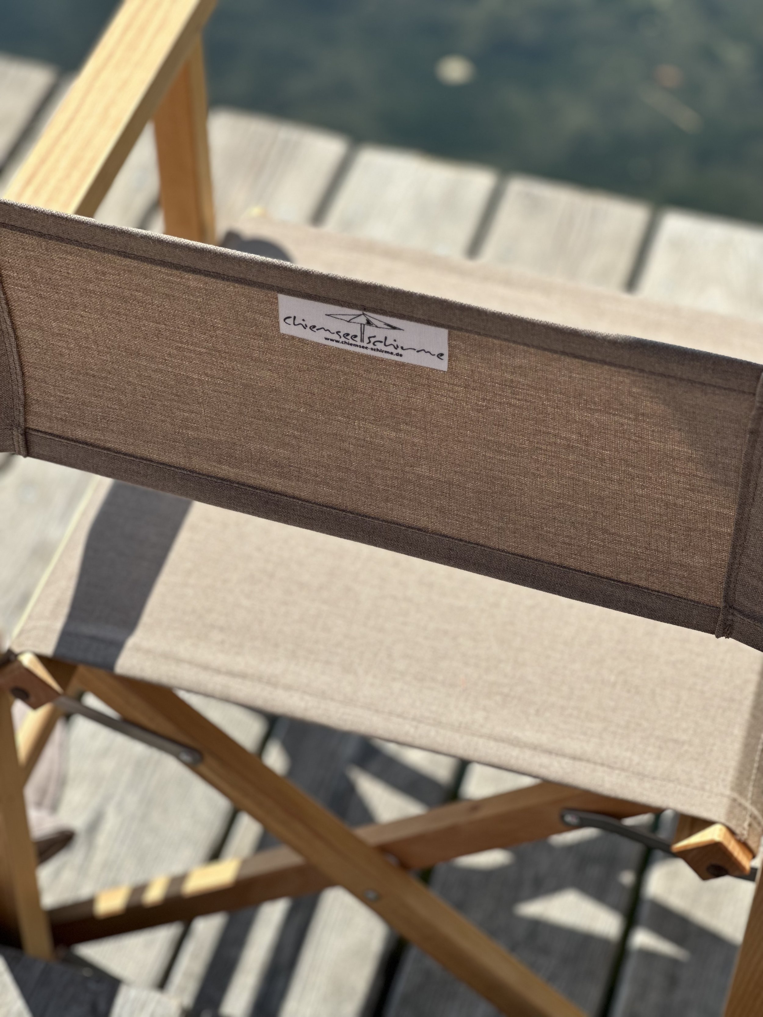 Nahaufnahme eines klappbaren Regiestuhls aus Holz und Stoff des Labels Chiemsee Schirme auf einem Holzdeck am Wasser.