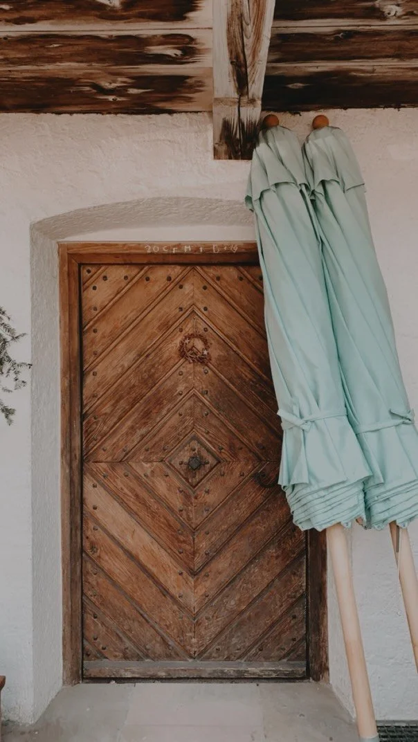 Ein hölzerner Tür in einer weißen Wand, darüber eine rustikale Holzbalken-Decke, neben der Tür stehen mehrere türkisfarbene Sonnenschirme.