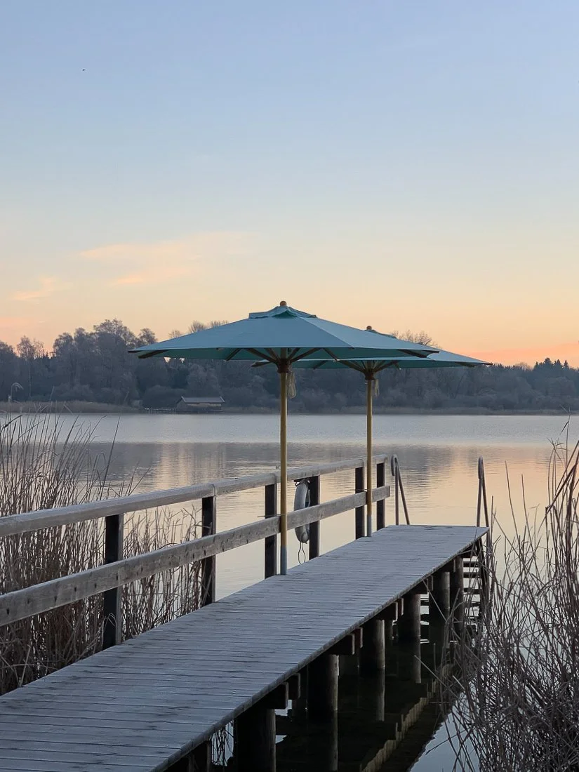 Ein Steg am Chiemsee mit einem aufgespannten Sonnenschirm von Chiemsee Schirme umgeben von Schilf, mit der Herreninsel im Hintergrund.