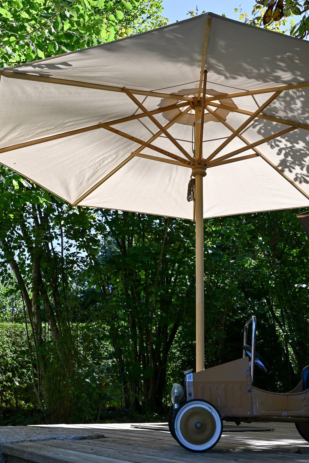 Großer weißer Sonnenschirm im Garten, darunter ein kleines Spielauto auf Holzterrasse, im Hintergrund viele grüne Bäume und Sträucher.