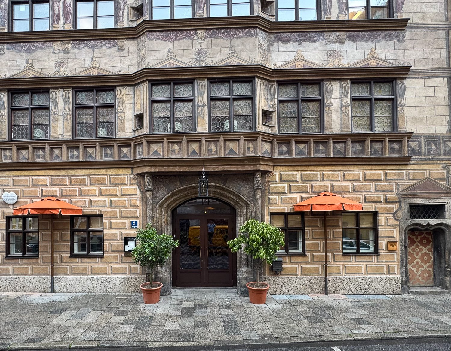 Historisches Gebäude mit steinverzierten Fassaden, mehreren Fenstern, zwei Terrassenschirmen mit Tischen, Pflanzen und Eingangstür in der Mitte.