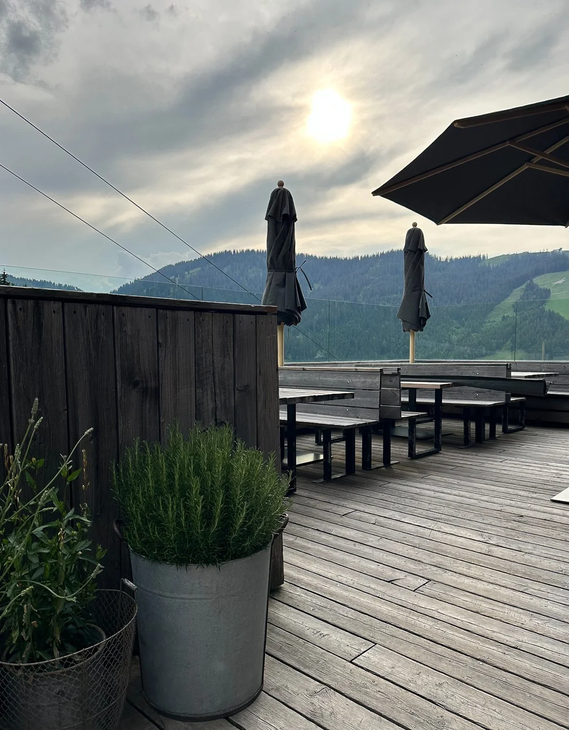 Ein ruhiger Terrassenausblick mit Holztisch, Sonnenschirmen, Bergland im Hintergrund, bewölkter Himmel, Pflanzen in Töpfen.