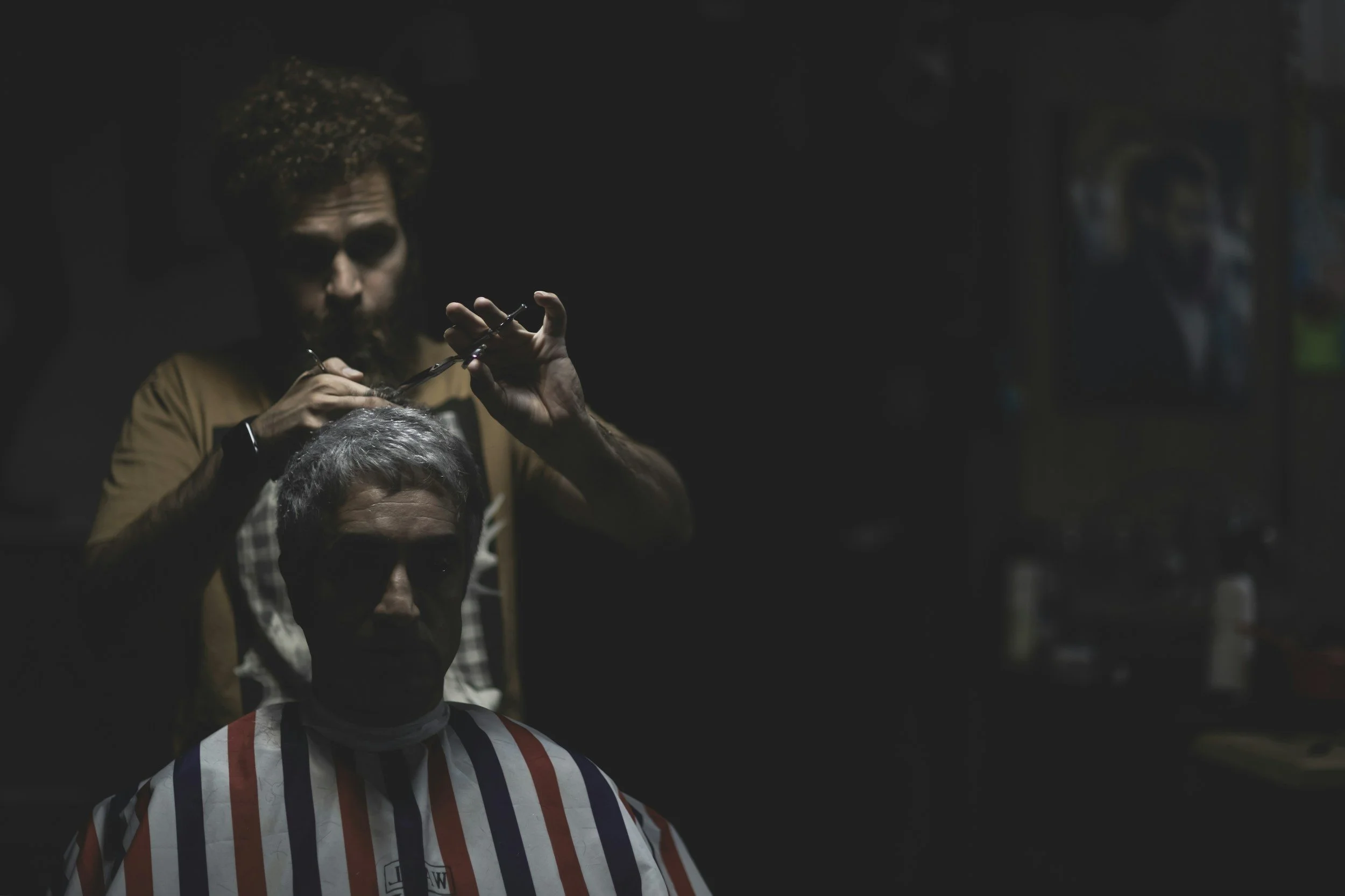 A barber giving a haircut to a woman with gray hair in a salon with dark lighting, using a pair of scissors while the woman wears a striped barber cape.