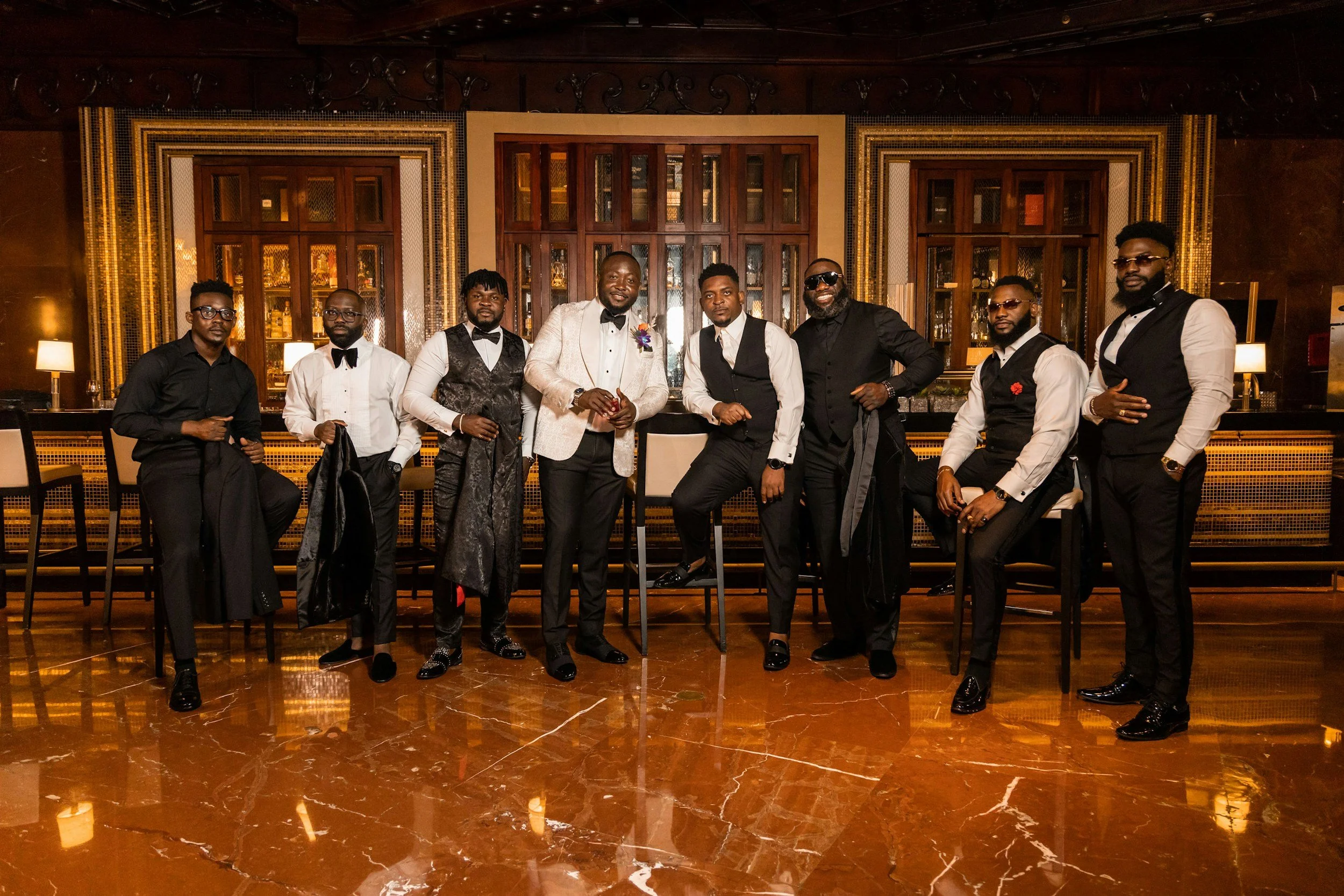 A group of nine men in formal attire posing in front of a bar with a wooden backdrop in an elegant setting.