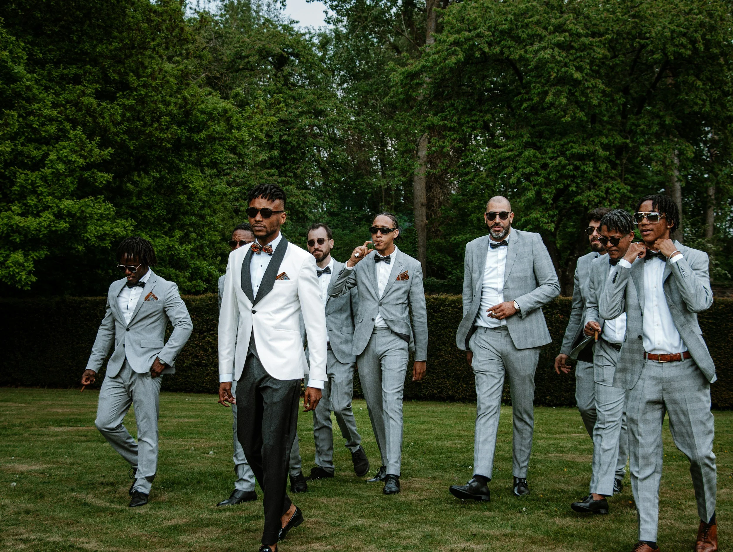 A group of men dressed in formal suits and sunglasses walking outdoors on a grassy area with tall trees in the background.