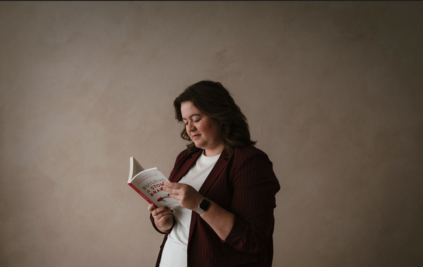 Woman with dark hair wearing a burgundy blazer and white t-shirt, reading a book titled 'Building a StoryBrand' against a plain beige wall.
