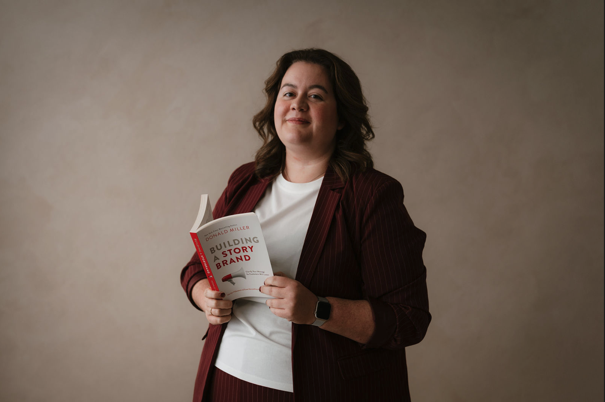 A woman with shoulder-length brown hair, wearing a white shirt and a maroon pinstripe blazer, holding a book titled "Building a Story Brand" by Donald Miller, standing against a plain beige background.