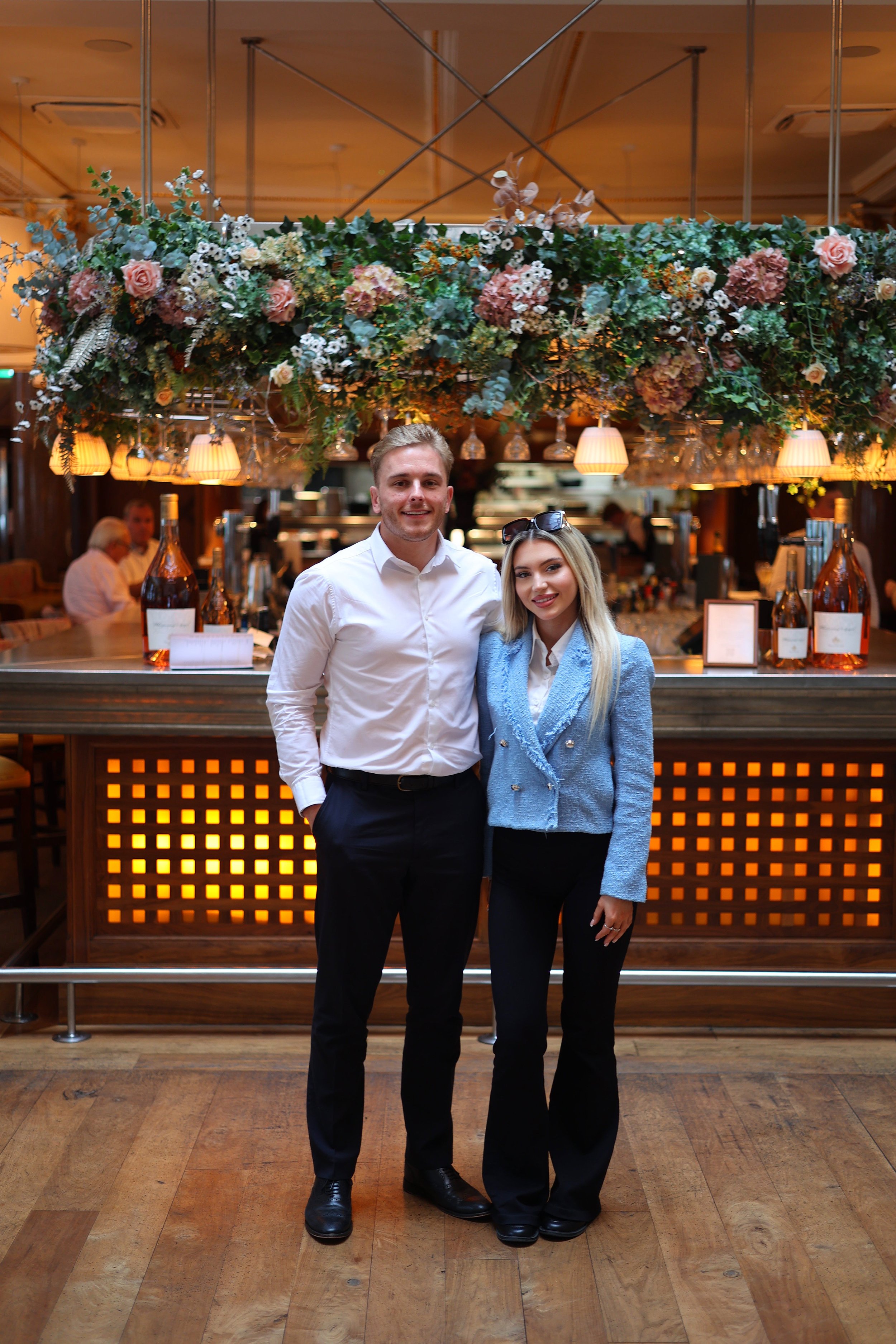 Luke and Rosie from excel accountants Liverpool standing together in a restaurant or bar, smiling at the camera, with a decorated floral display above them.