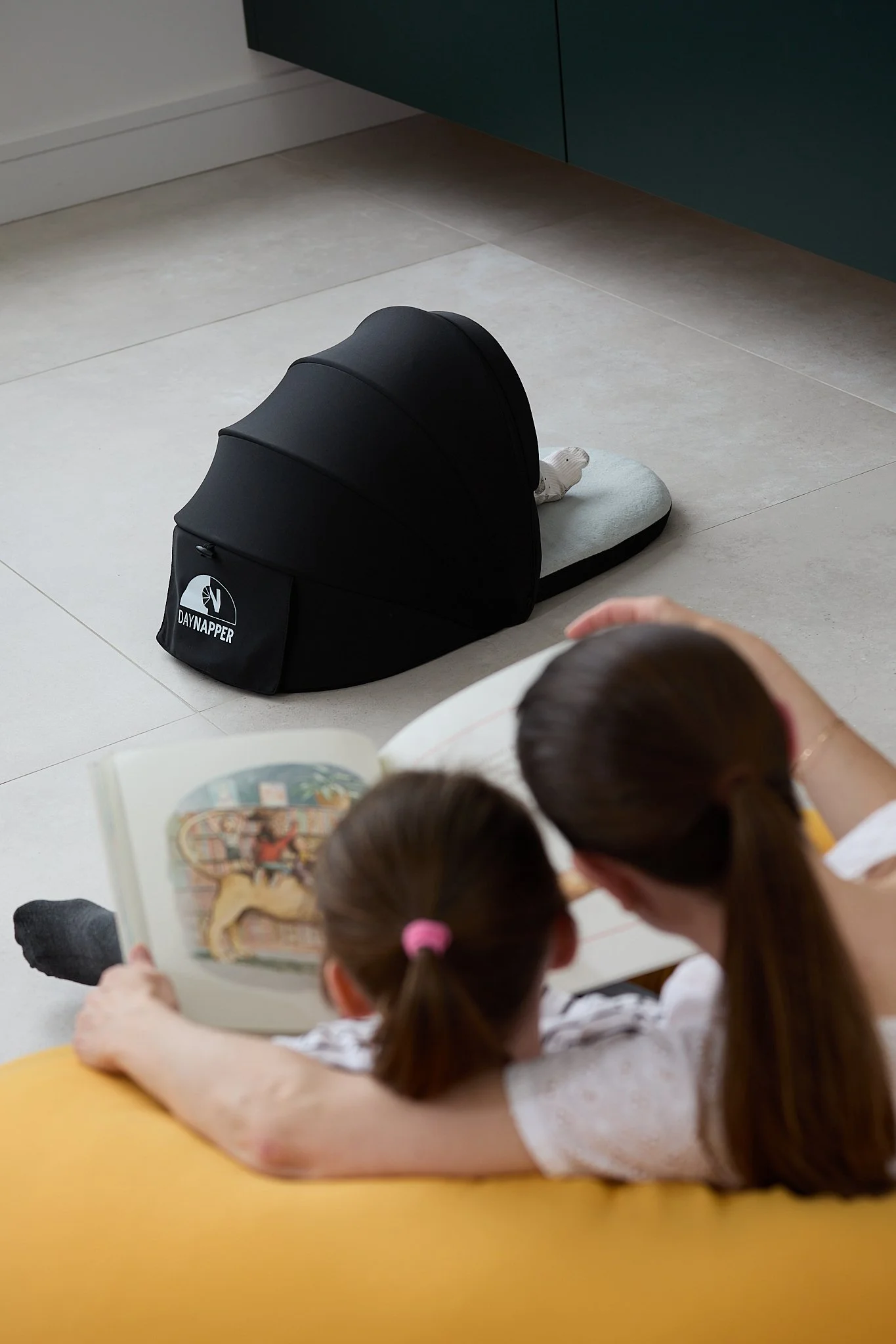 A mother and girl reading a colourful picture book on a yellow sofa with a baby lying in DayNapper on the tiled floor.