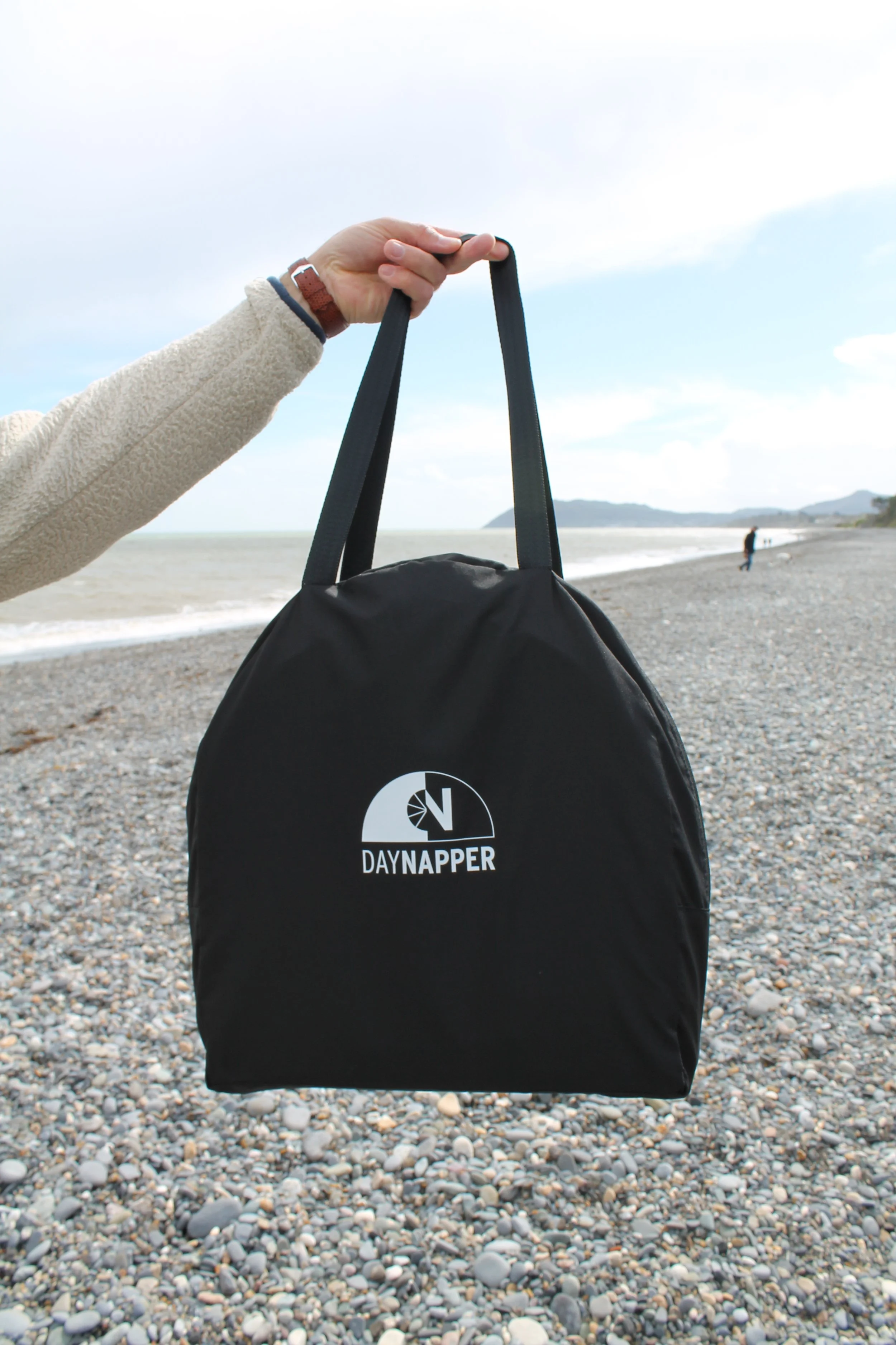 A person holding a black DAYNAPPER bag by its handles on a pebble beach, with ocean waves and a partly cloudy sky in the background.