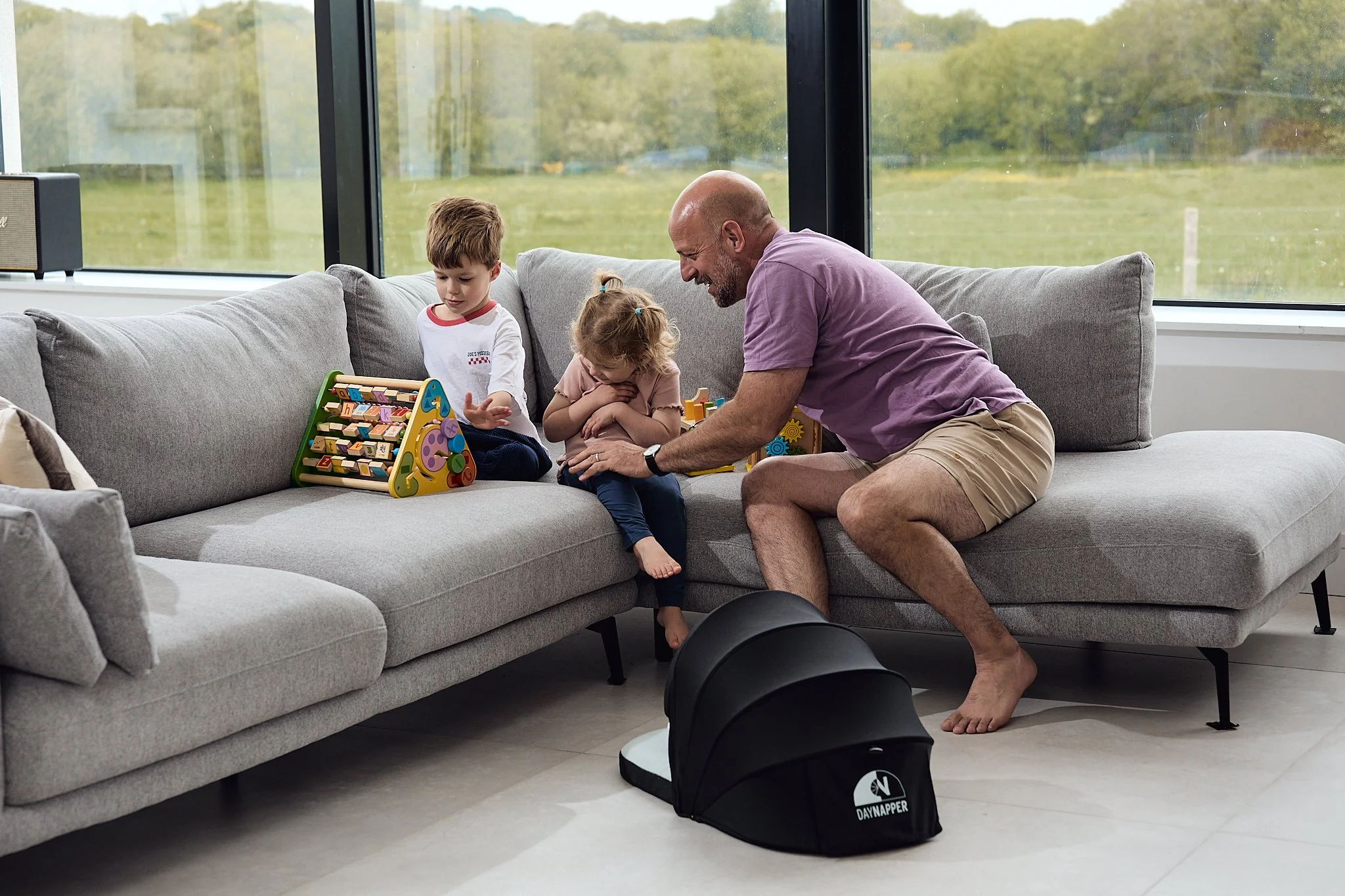 A father and two children playing with educational toys on a grey sofa in a sunlit living room with large windows overlooking a green outdoor landscape and baby in a DayNapper on the floor.