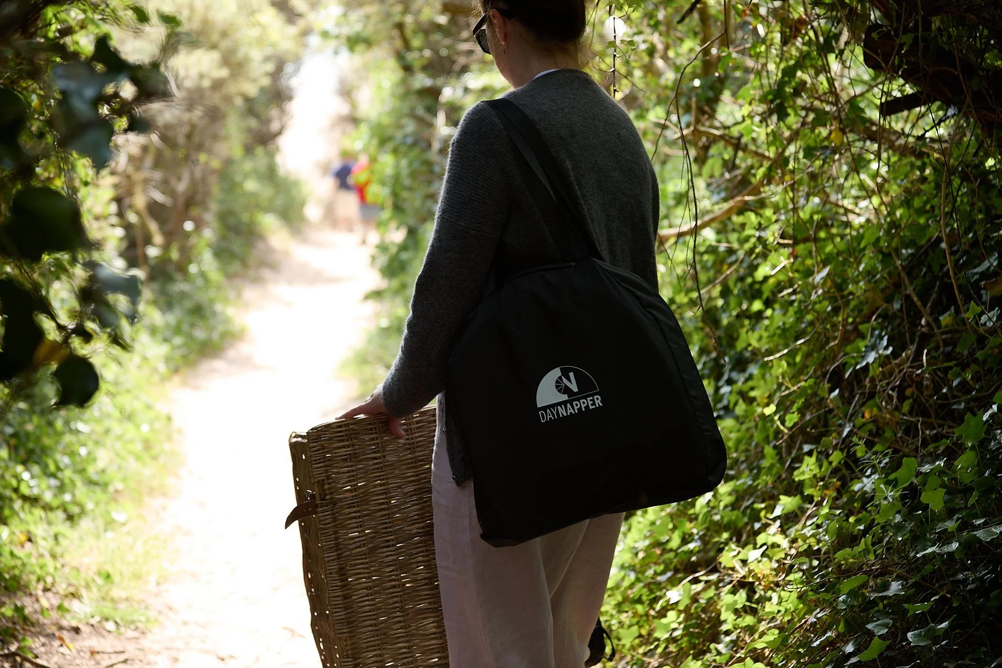 A mother walking through a lush, green forest trail with a wicker basket in her hand, wearing a grey sweater and carrying a DayNapper in the portable storage and carry bag.